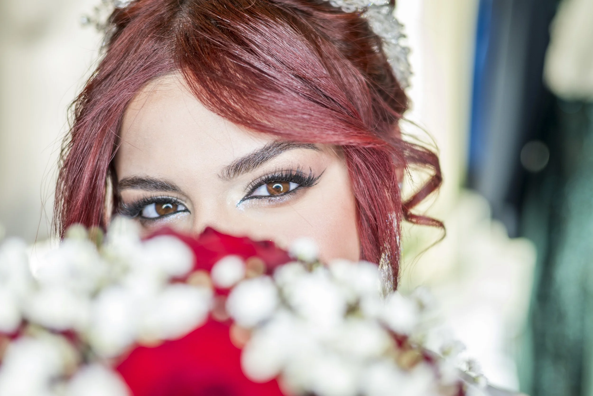 Close-up of a woman's face with brown eyes, red hair styled with loose curls, and makeup with eyeliner and mascara. She is partially obscured by white and red flowers in the foreground.