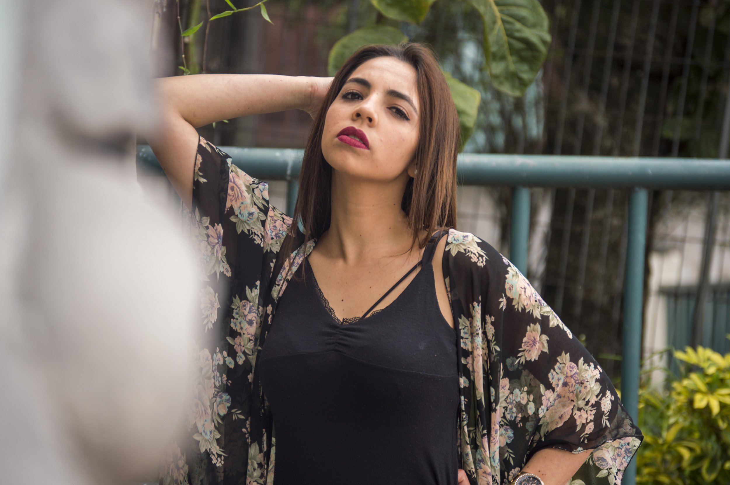 A woman with shoulder-length brown hair, wearing a black top with a floral sheer cover-up, stands outdoors with her hand resting on her head, looking confidently at the camera. There is greenery and a metal fence in the background.