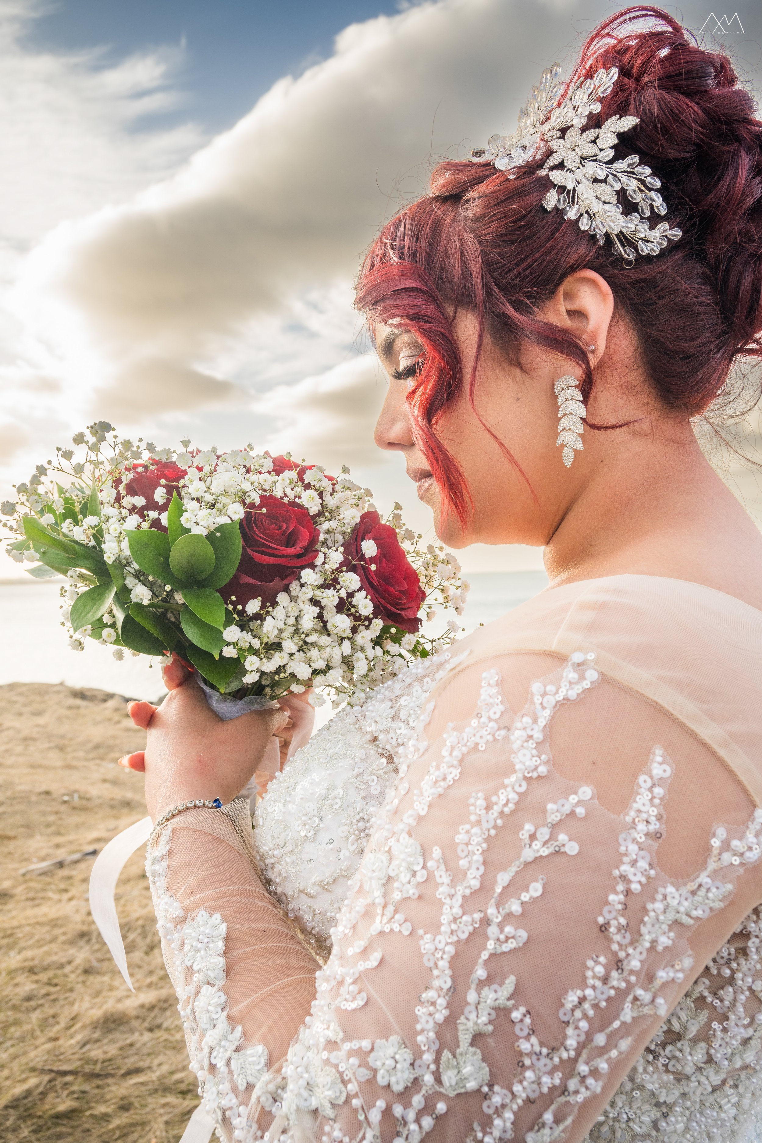 A bride with red hair styled in an elegant updo, holding a bouquet of red roses and white baby's breath, stands outdoors near the water with clouds in the sky.