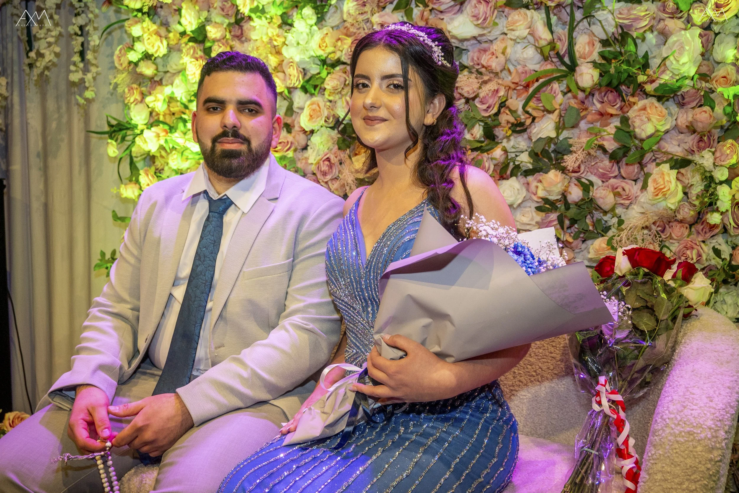 A couple dressed in formal attire sitting in front of a floral backdrop, with the woman holding a bouquet of flowers.