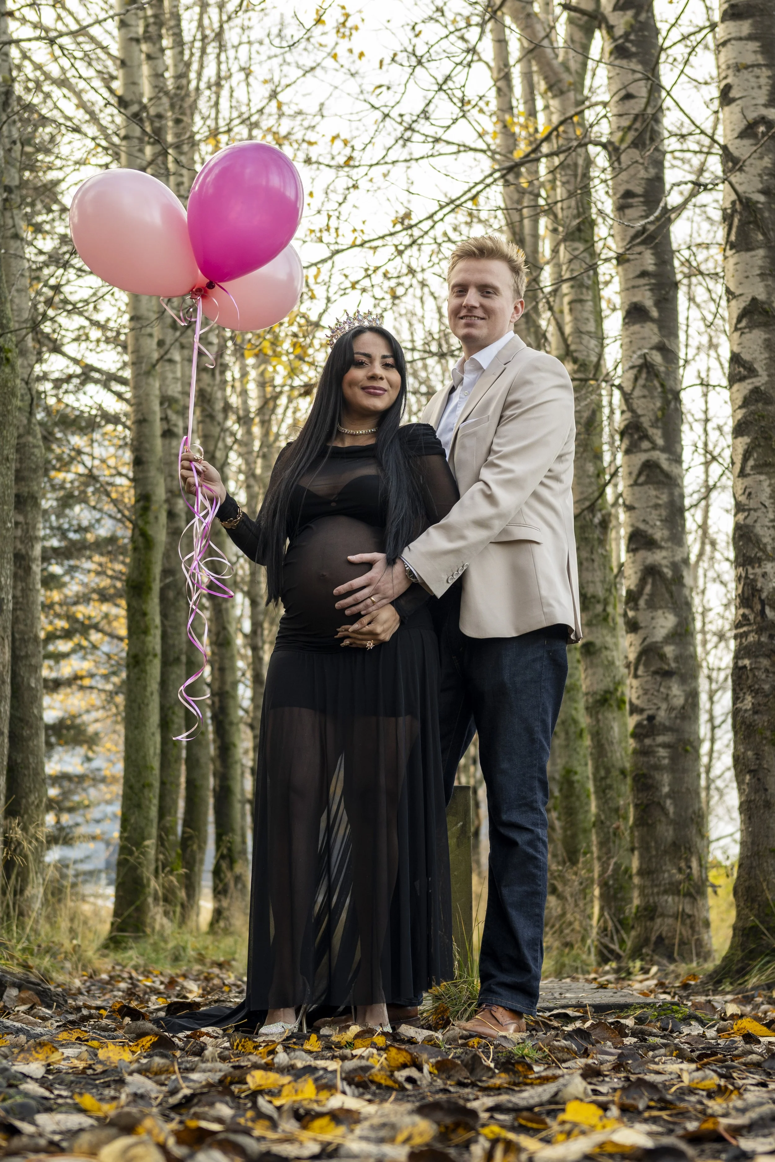 A pregnant woman wearing a black dress and tiara, holding pink balloons, is standing outdoors in a wooded area with a man in a beige blazer, in a sunny forest setting with fallen leaves.