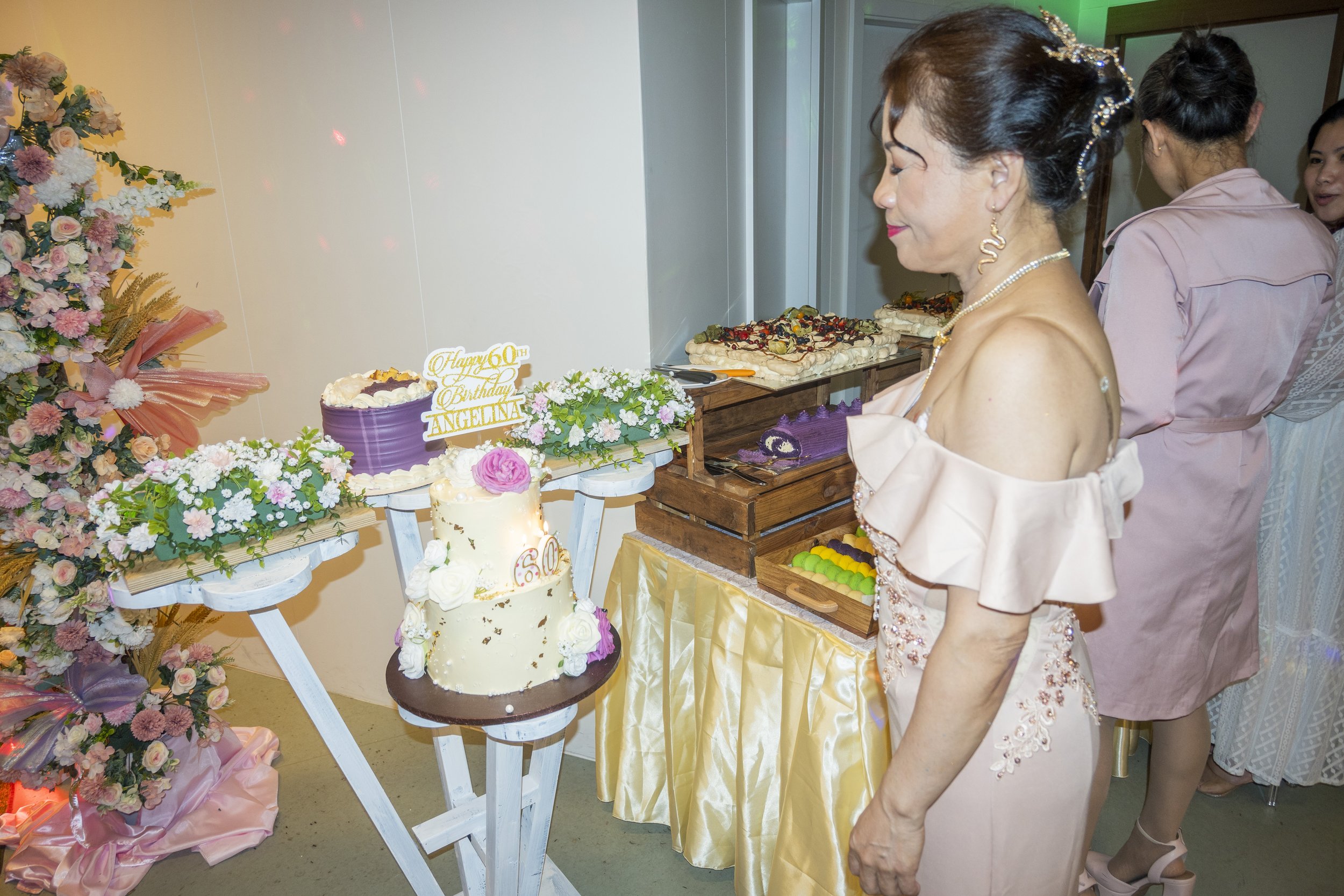 Woman in pink dress celebrating birthday with a decorated cake, surrounded by floral arrangements and others serving food.