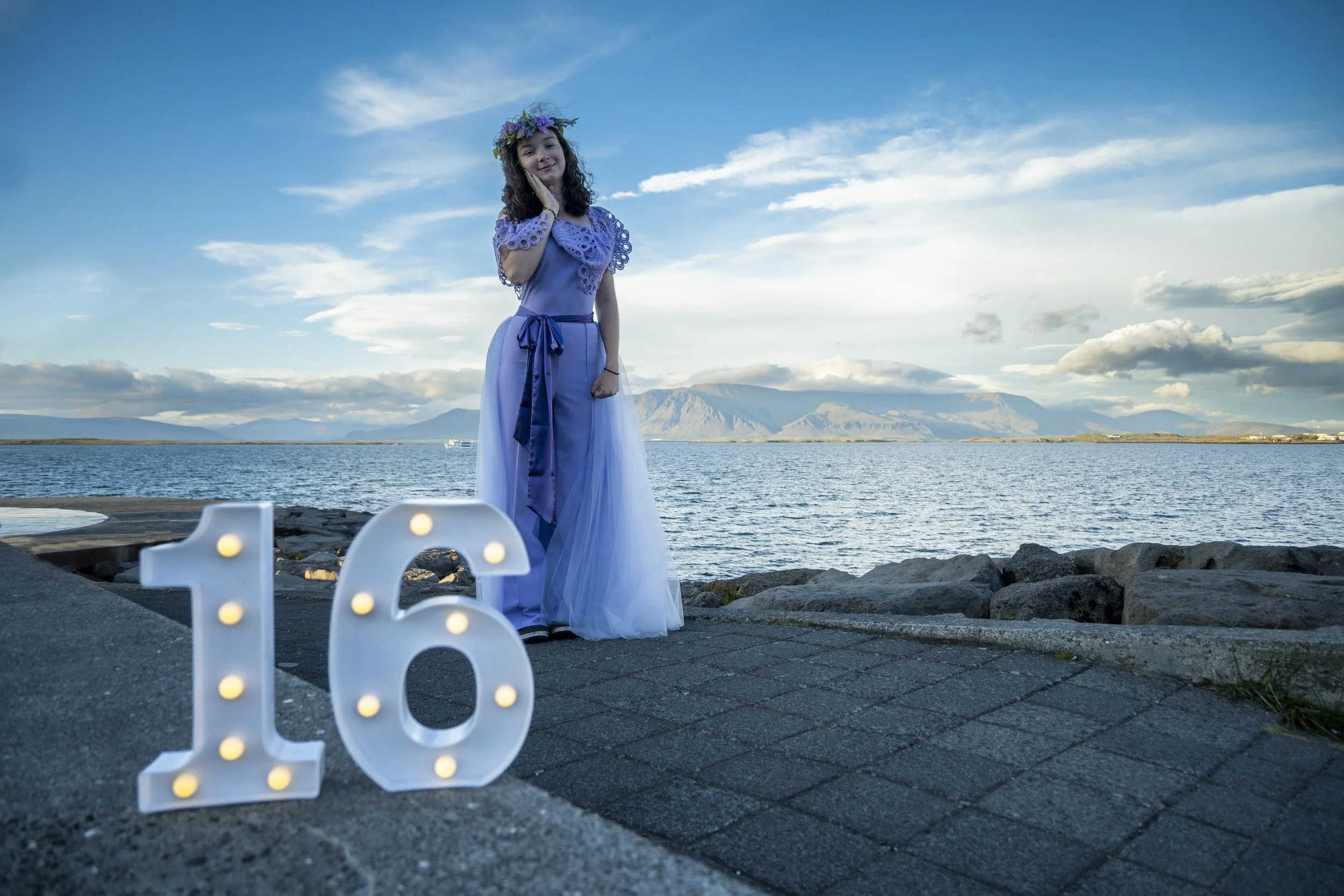 A girl standing by a lakeside wearing a blue dress with a flower crown, next to illuminated number 16, during sunset or late afternoon, with mountains in the background.