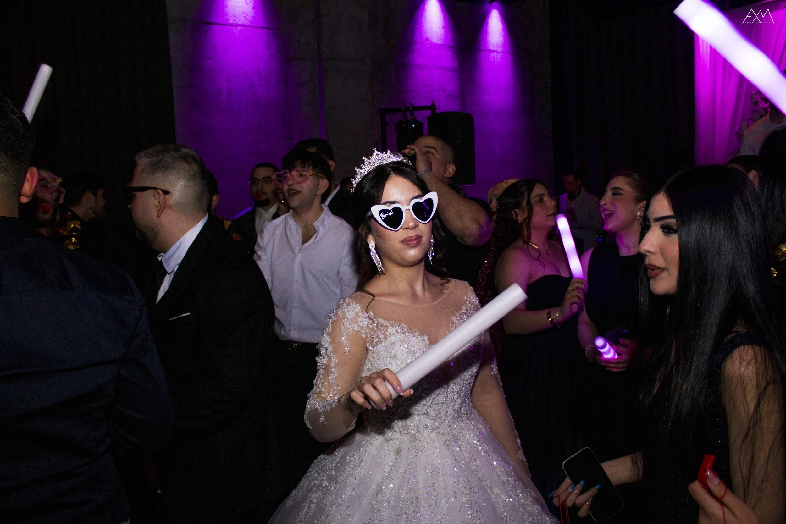 A woman dressed in a wedding gown holding a glow stick at a celebration party, surrounded by other people in formal attire with colorful lighting.