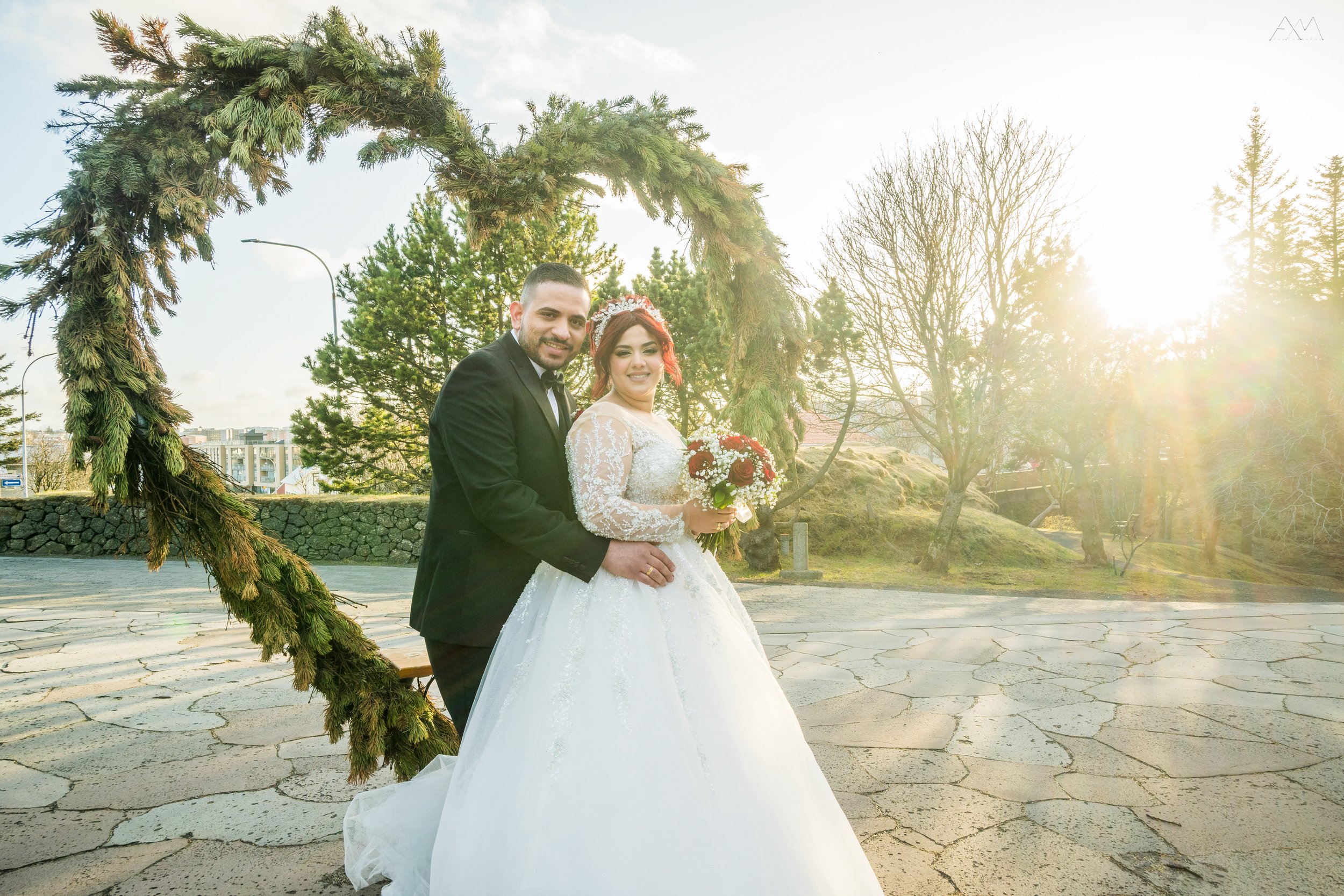 A newlywed couple in wedding attire standing outdoors under a circular greenery arch, with sunlight shining through trees in the background, the bride holding a bouquet of flowers.