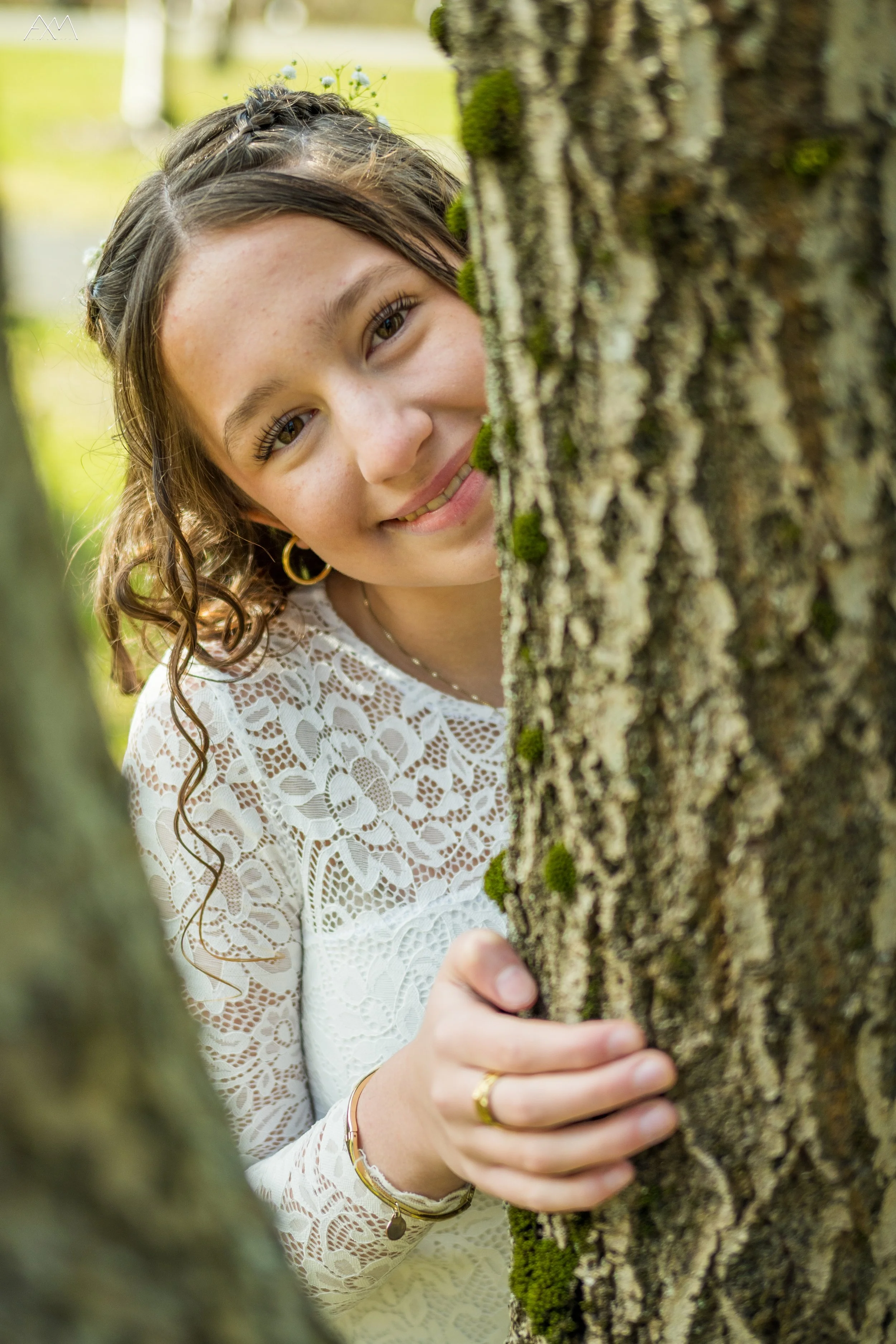 A young woman with curly brown hair, wearing gold hoop earrings, gold bracelets, and a white lace top, peeks from behind a tree trunk with a happy expression.