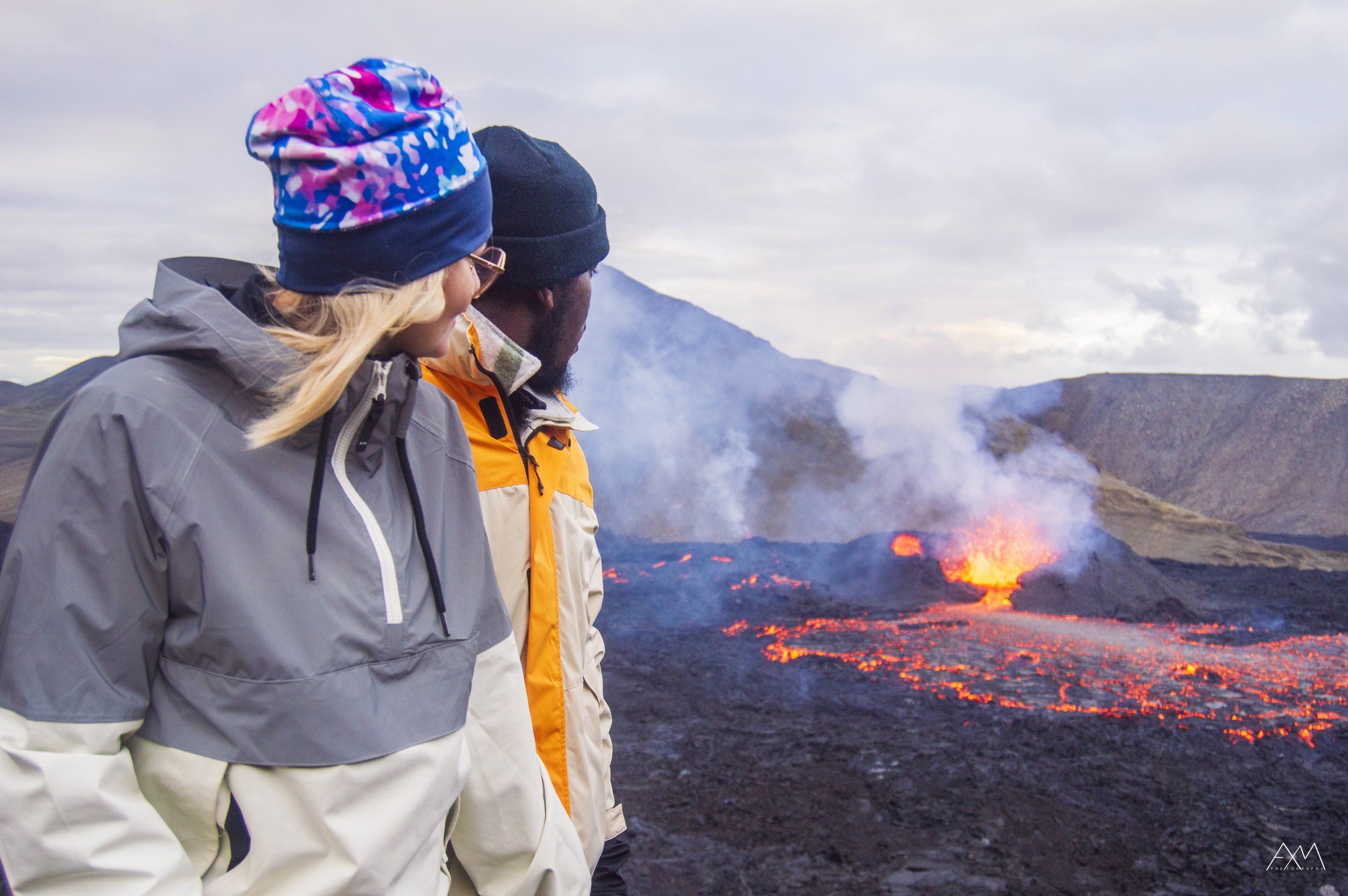 Two people in outdoor jackets and beanies observing an erupting volcano with flowing lava and smoke.