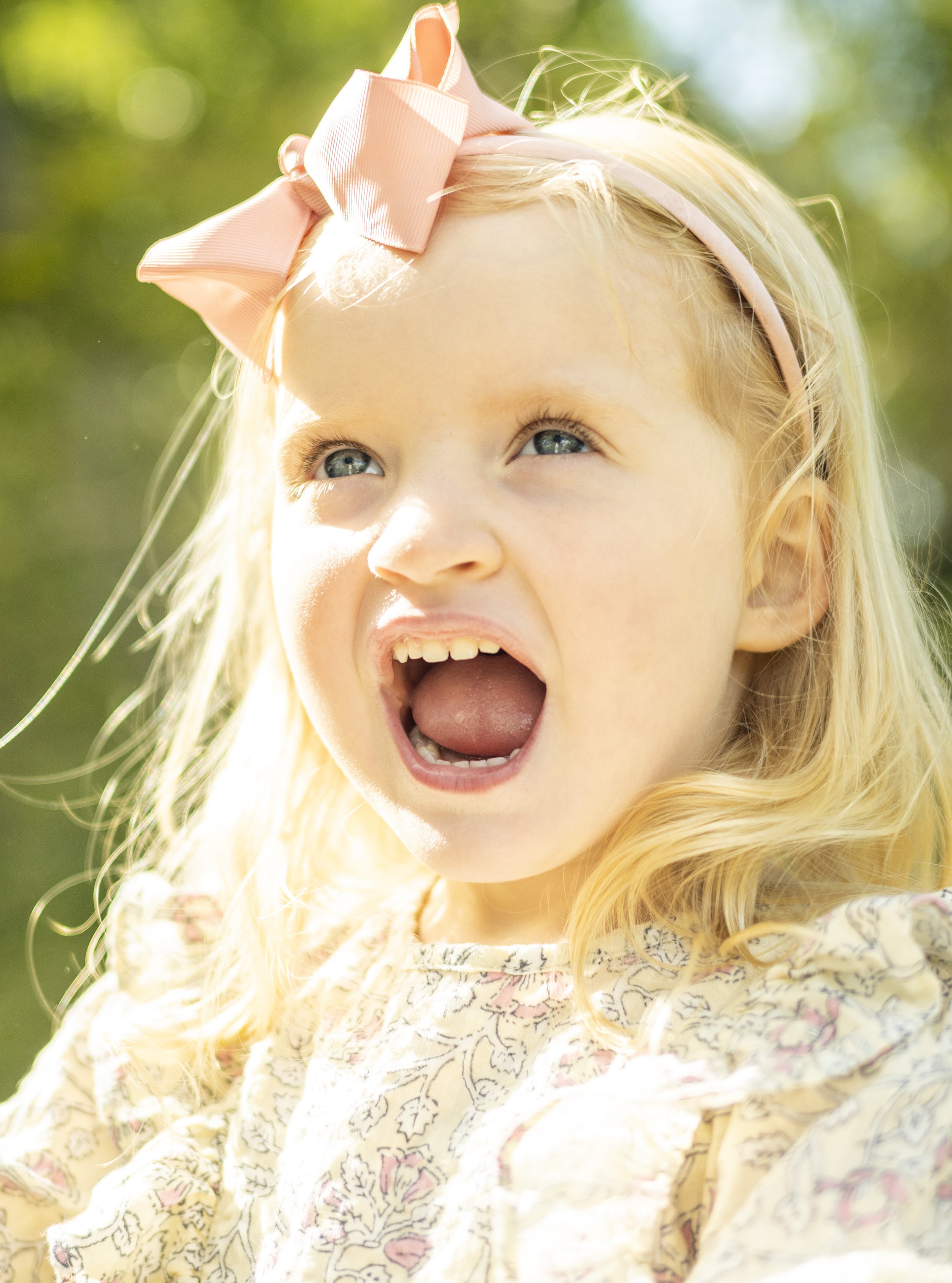Young girl with blonde hair, wearing a pink bow headband, expressing surprise or anger outdoors in bright sunlight.