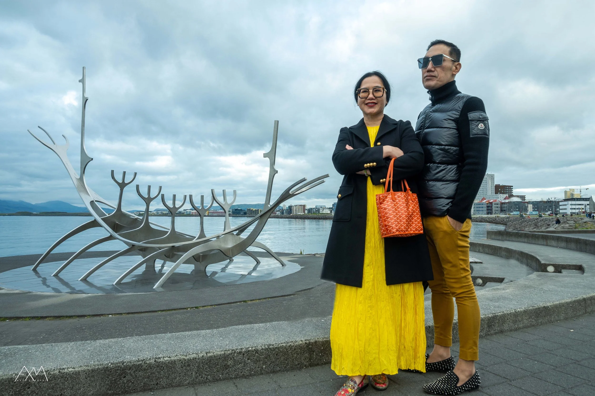 A woman in a yellow dress and a black coat with glasses and a red handbag, and a man in a black vest and mustard pants with sunglasses, standing near a metal sculpture shaped like a ship on a waterfront, with city buildings and cloudy sky in the back