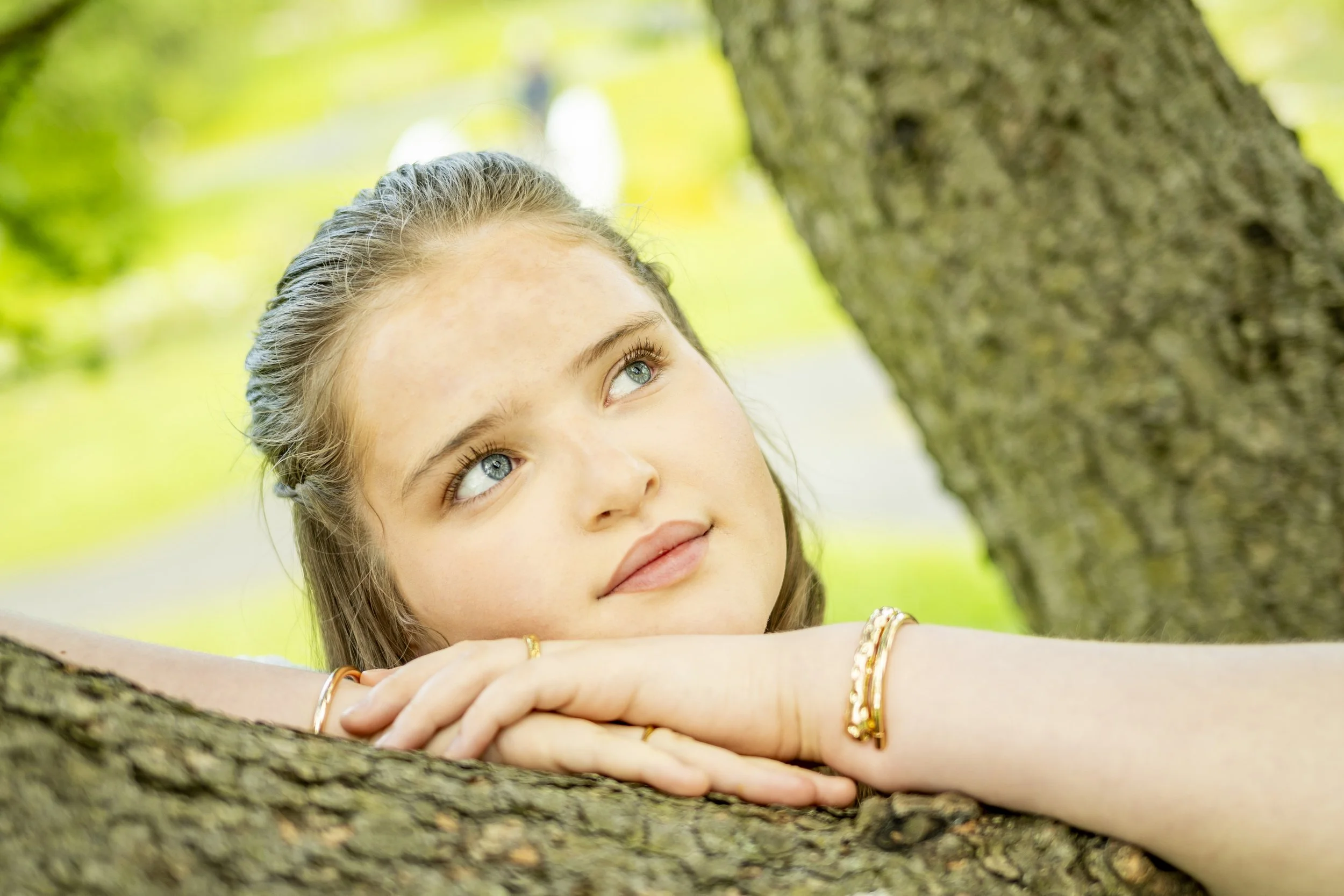 A young woman with blue eyes and light brown hair rests her arms on a tree branch, looking up through the branches and leaves in a lush, green outdoor setting.