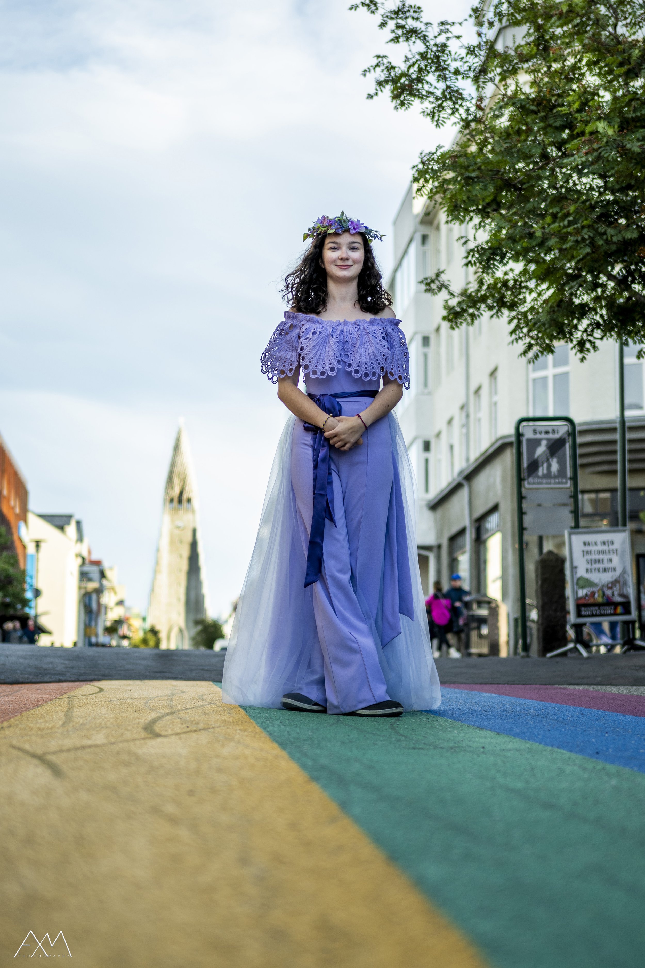 Young woman standing on a rainbow-street in Iceland crosswalk during daylight, dressed in a purple gown with intricate lace details and a flower crown, with city buildings and a church steeple in the background.