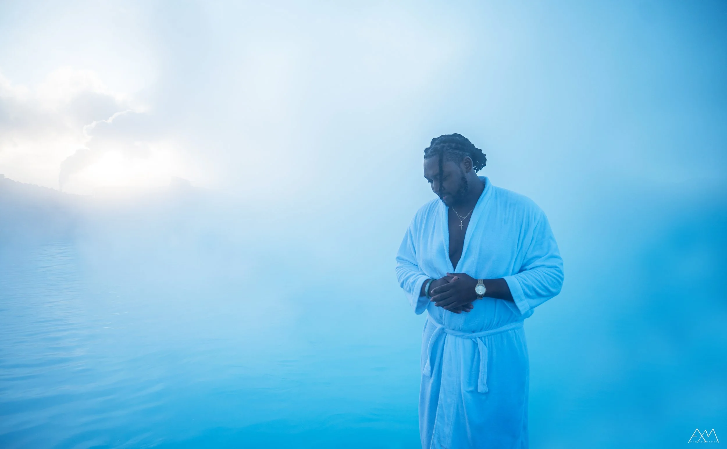 A man dressed in a white bathrobe standing with hands clasped in front of a foggy, blue-lit body of water with mist and clouds in the sky.
