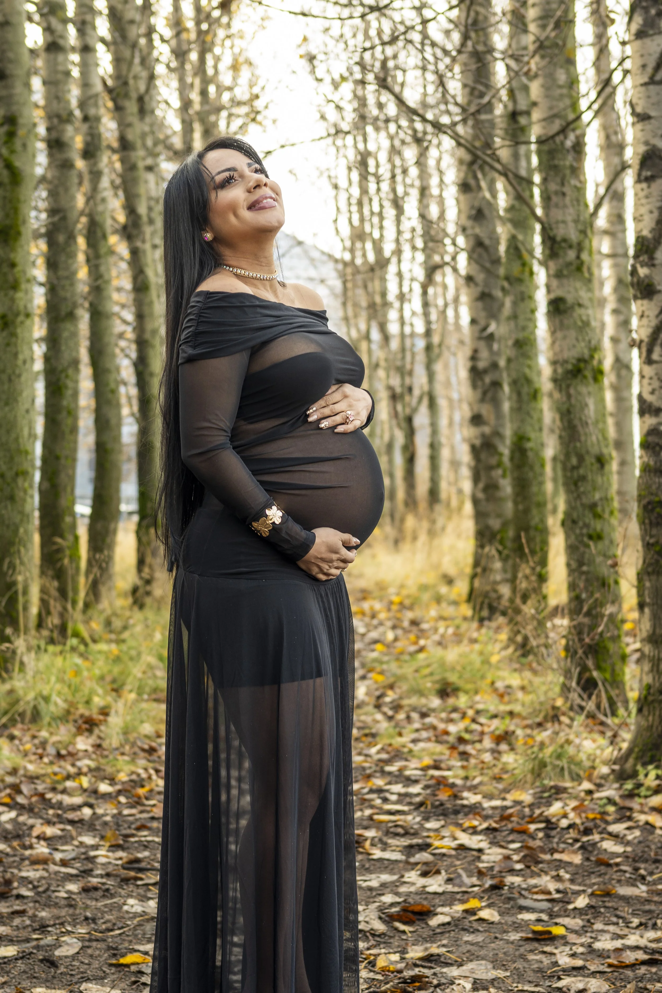 A pregnant woman with long dark hair standing in a forest with many trees and fallen leaves, wearing a black off-shoulder dress with sheer fabric, jewelry, and smiling while looking upward.