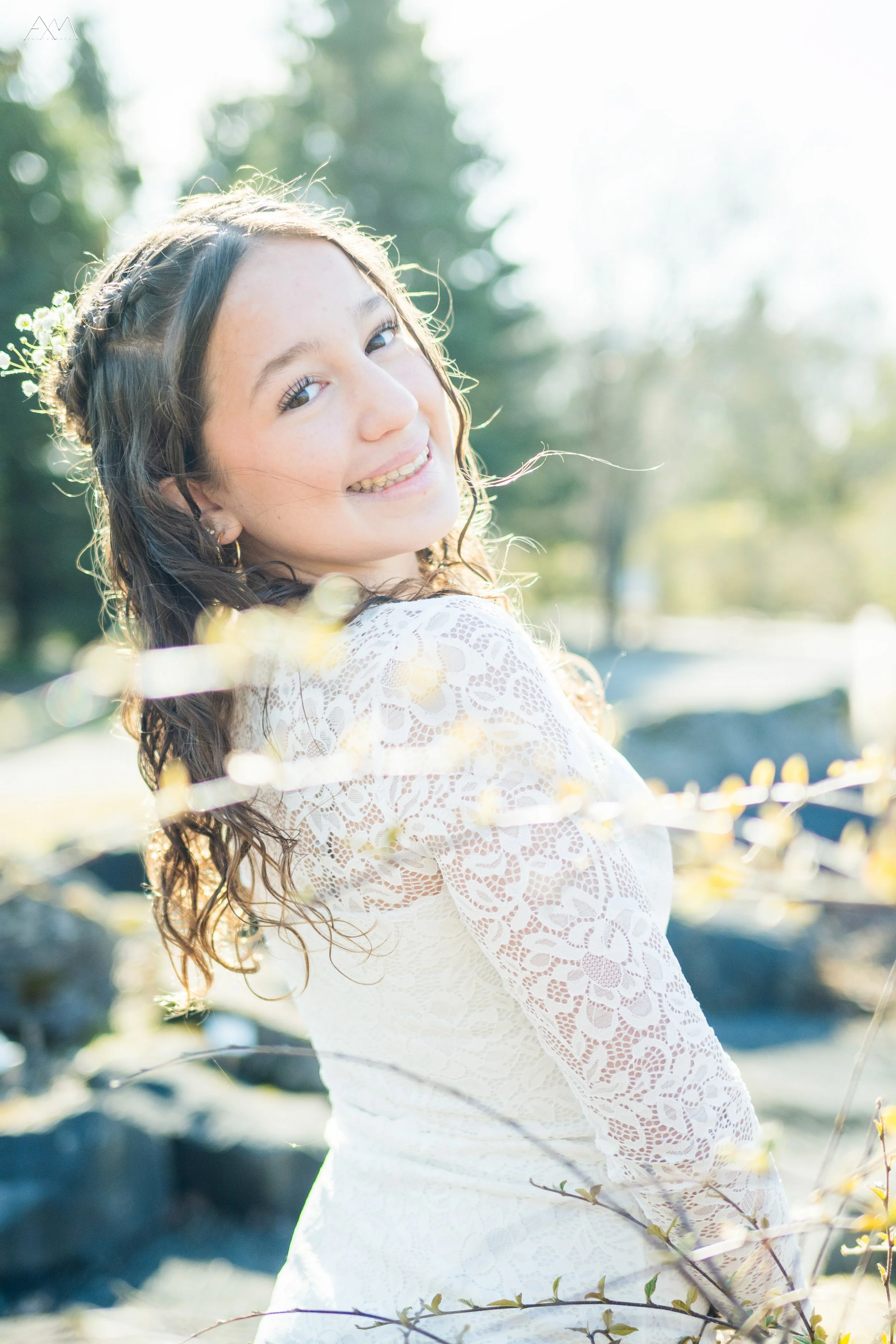 Smiling young woman with wavy hair outdoors in sunlight, wearing a white lace dress, surrounded by trees and branches.