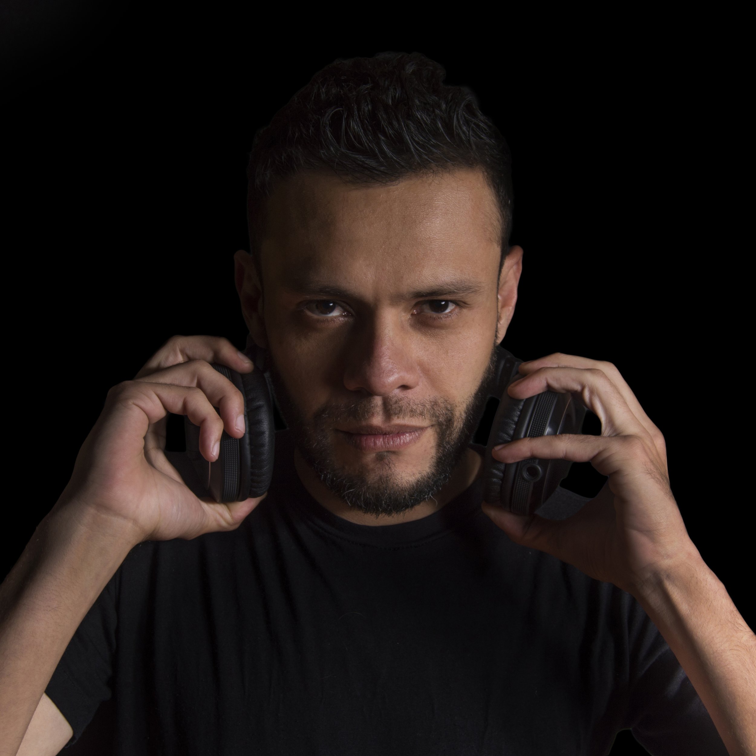 Man with dark hair and beard holding headphones near his ears against a black background.