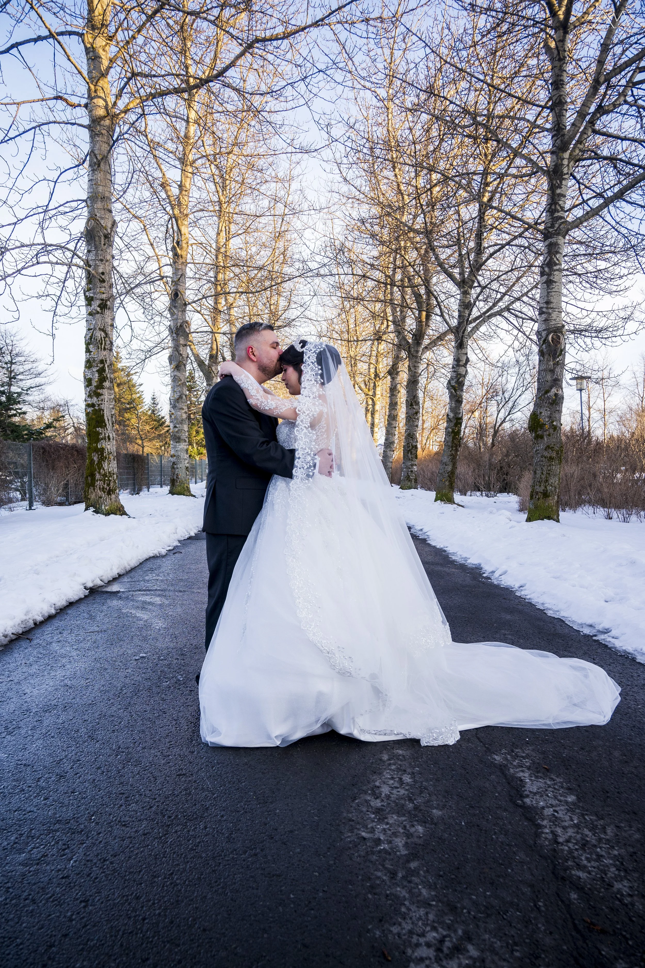 A bride and groom embracing and kissing outdoors during winter, with bare trees and a snowy ground.