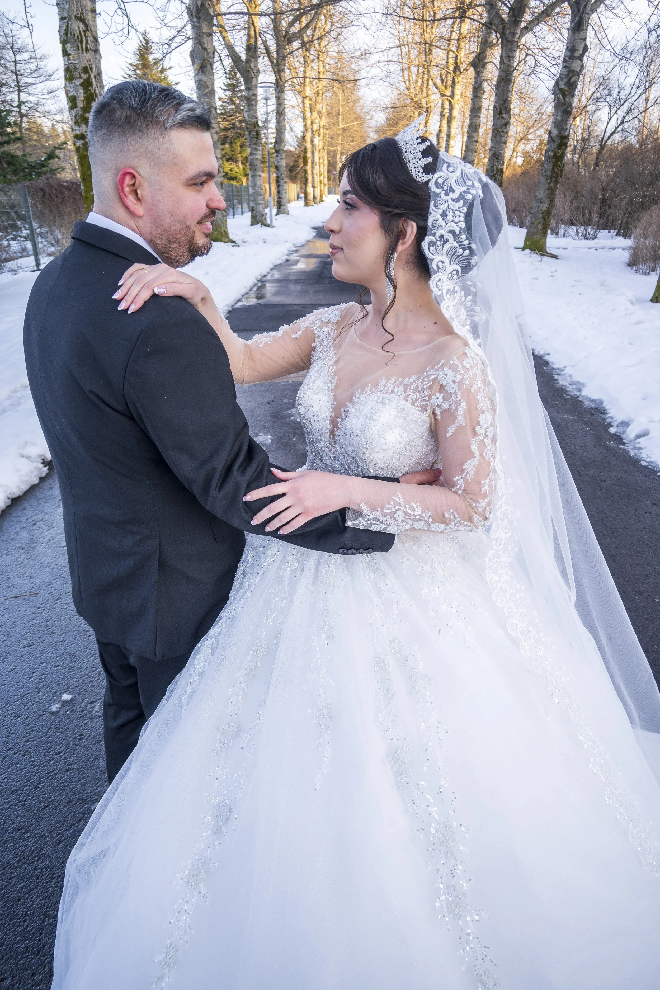 A bride and groom standing close together outdoors in a snowy environment, looking into each other's eyes, with trees in the background. The bride wears a white wedding gown with lace and a veil, and the groom is in a black suit and tie.