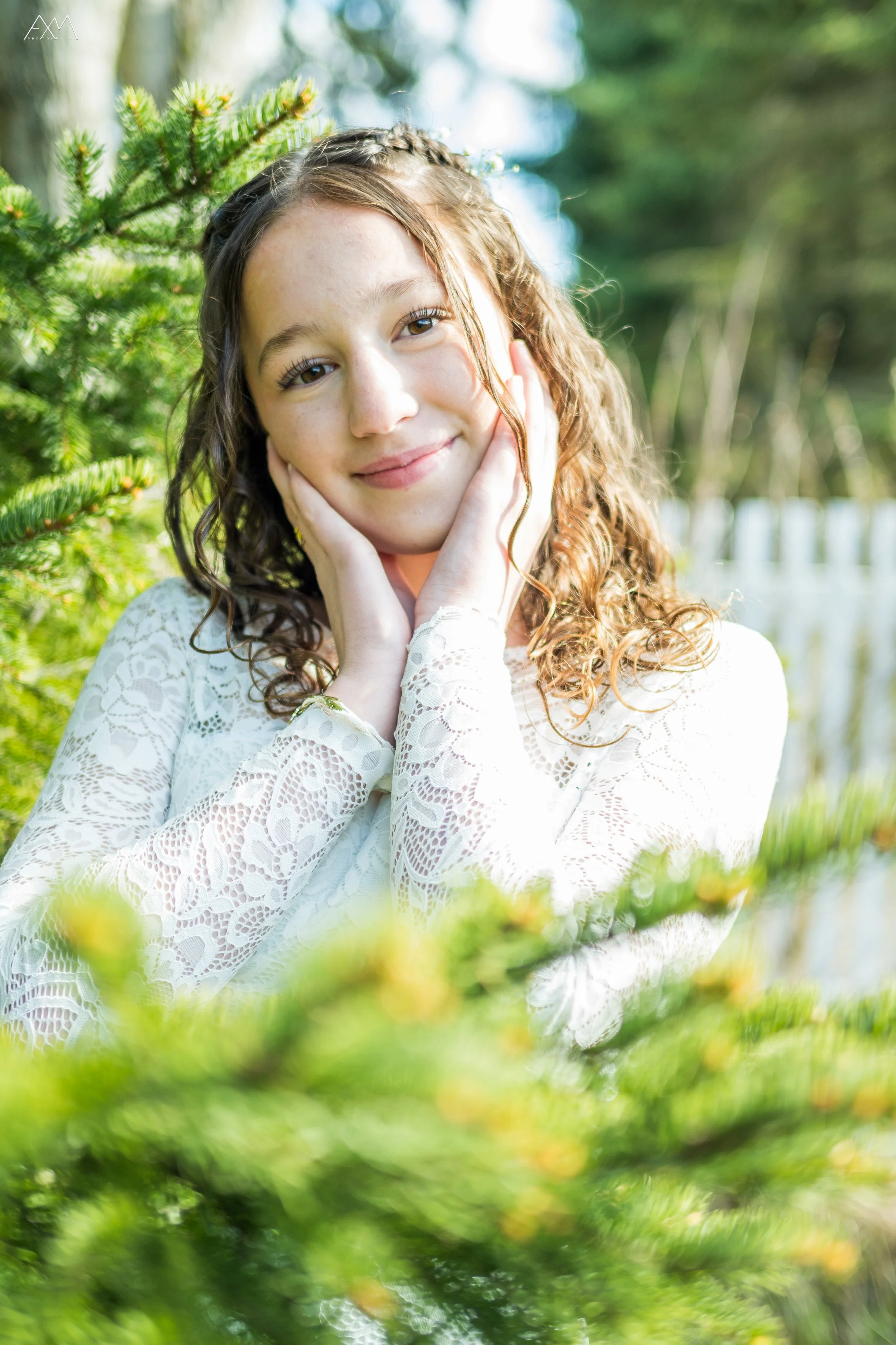 A young girl with curly brown hair gently holding her face with her hands, smiling softly, outdoors with green trees and sunlight in the background.