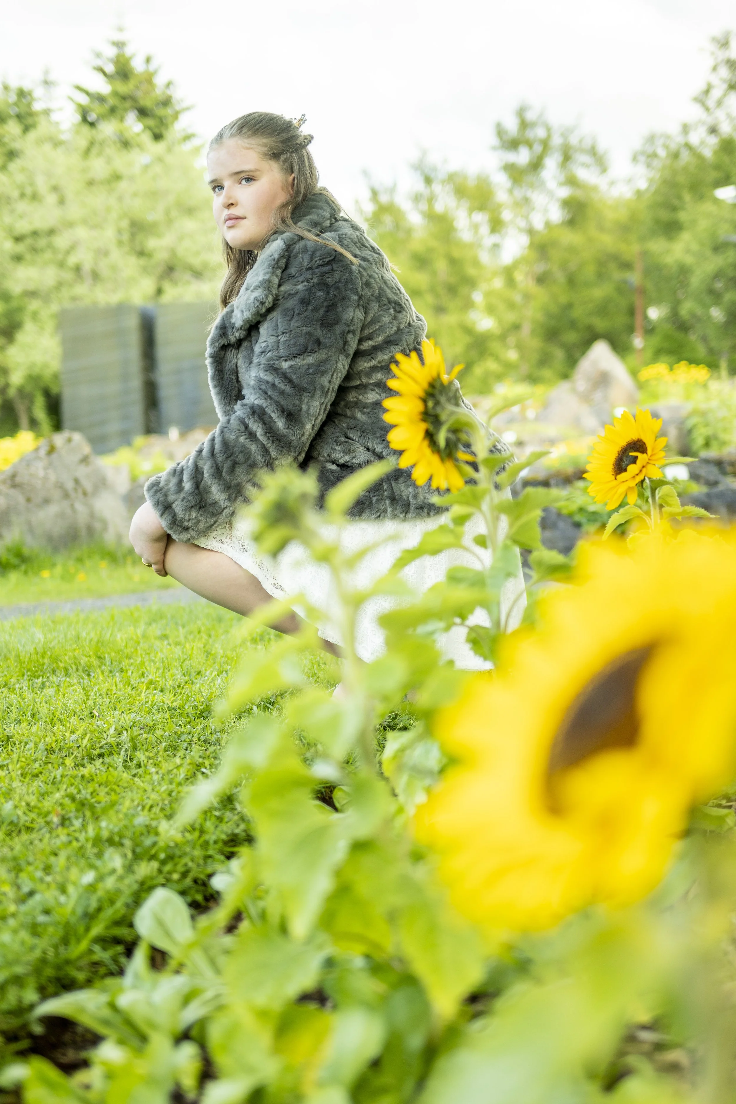 A young girl with brown hair and blue eyes wearing a fuzzy gray coat, sitting outdoors in a garden with yellow flowers and green trees, looking to the side.