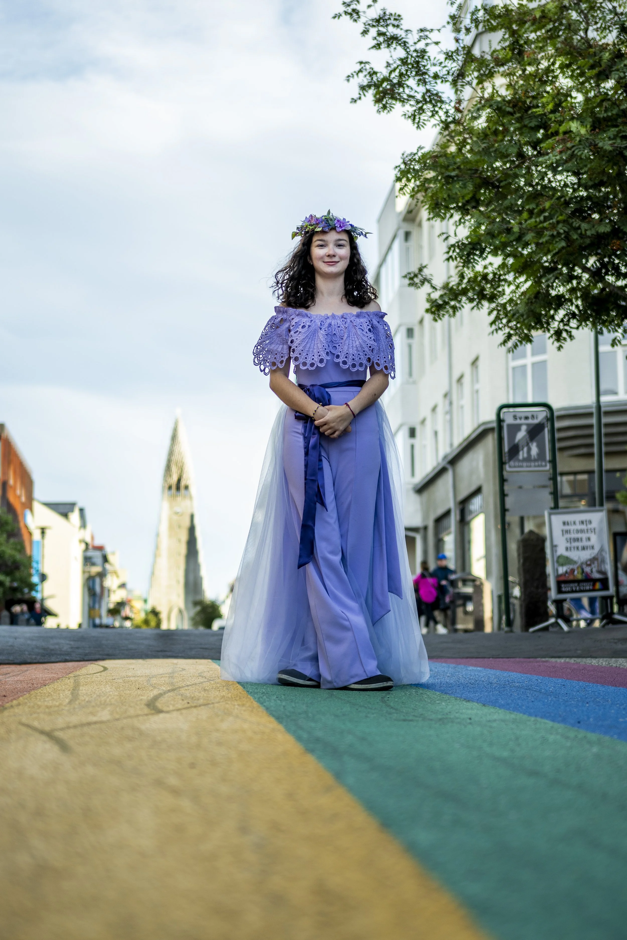 A young woman with dark, curly hair and a flower crown, dressed in a purple lace top and a long, flowing purple skirt, standing on a rainbow crosswalk in an urban area with buildings and a church steeple in the background.
