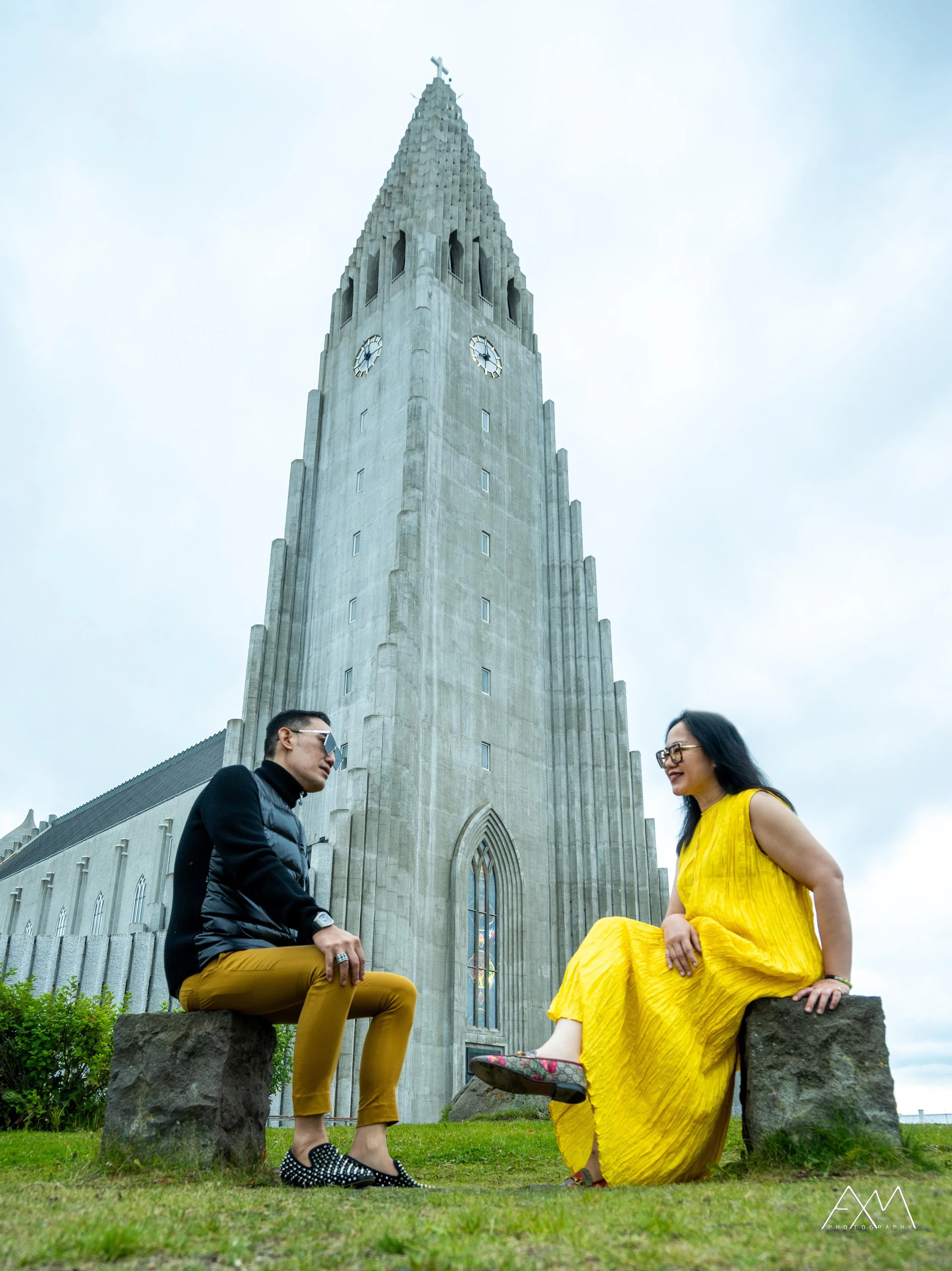 A man and woman sitting on stone benches in front of Hallgrímskirkja church in Reykjavik, Iceland. The man is wearing glasses, a black jacket, and yellow pants, while the woman is dressed in a flowing yellow dress and sandals. The gray church tower w