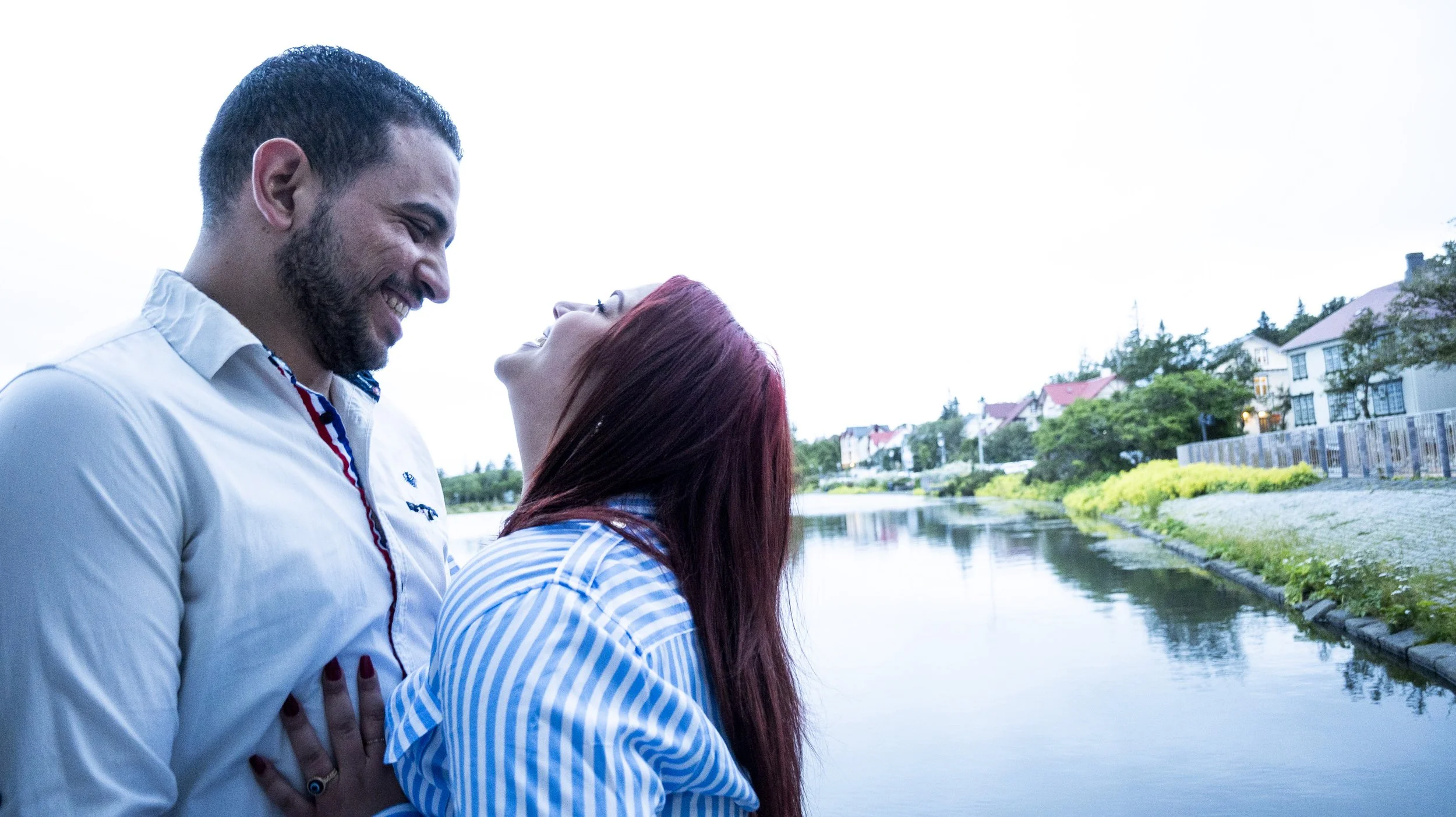 A couple smiling and laughing beside a river with houses and greenery in the background.
