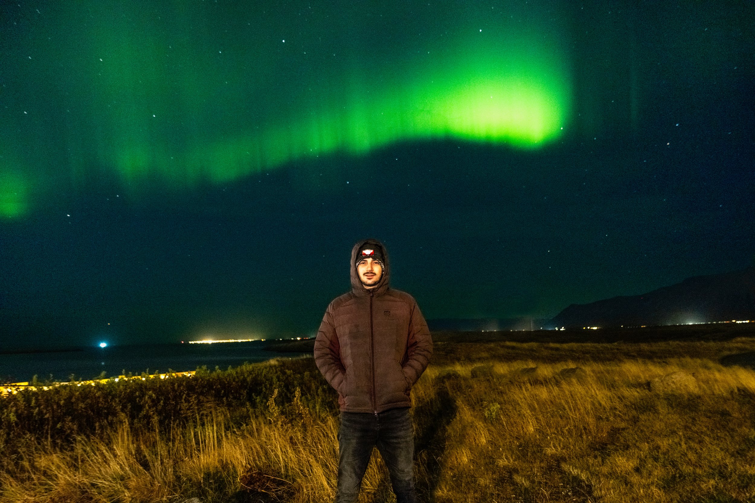 A man wearing a brown jacket and dark pants standing on a grassy field at night with the Northern Lights glowing green in the sky behind him.