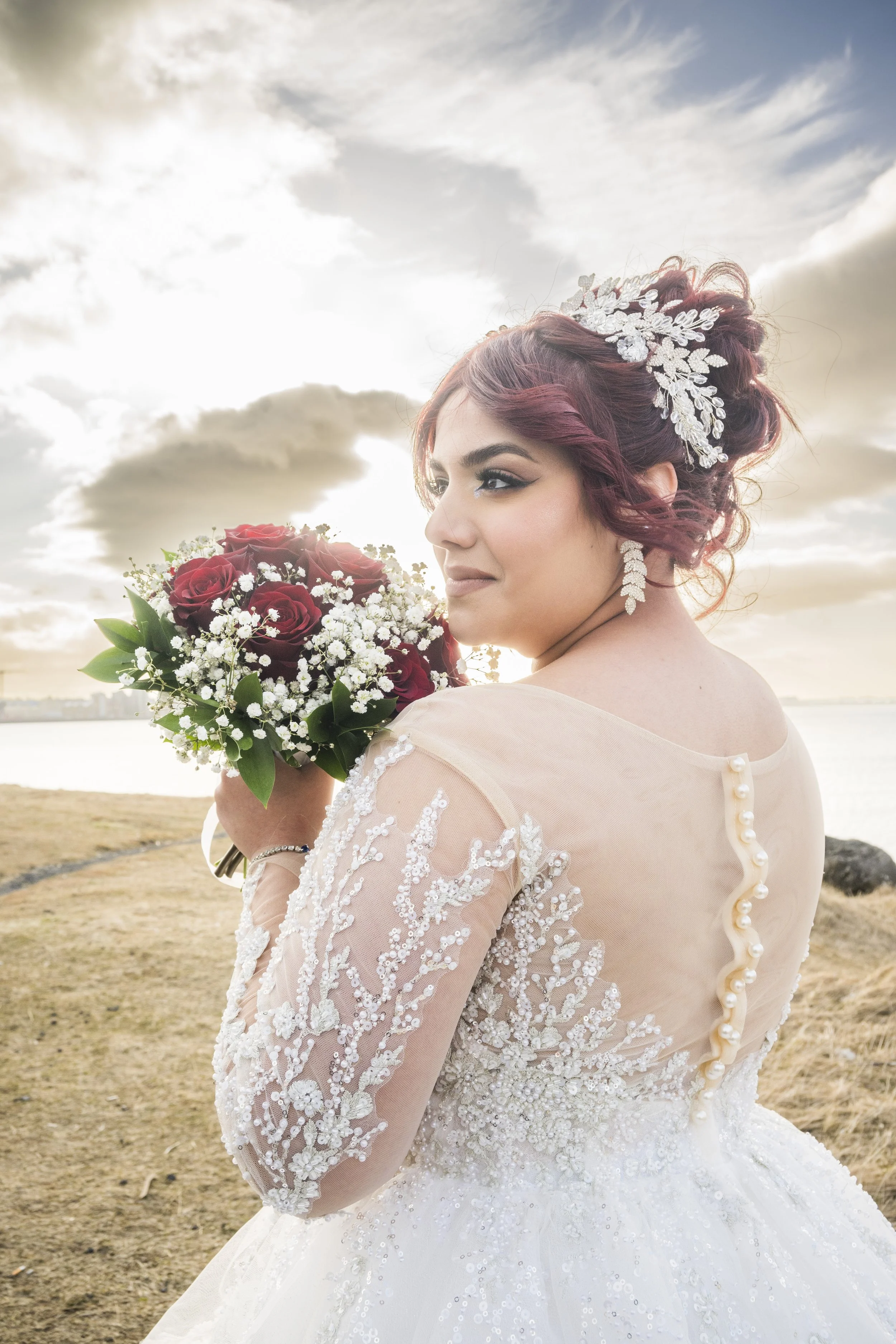 A bride with burgundy hair styled in loose curls, wearing a detailed lace wedding gown with pearl buttons on the back, holding a bouquet of red roses and white baby's breath, outdoors with cloudy sky and water in the background.
