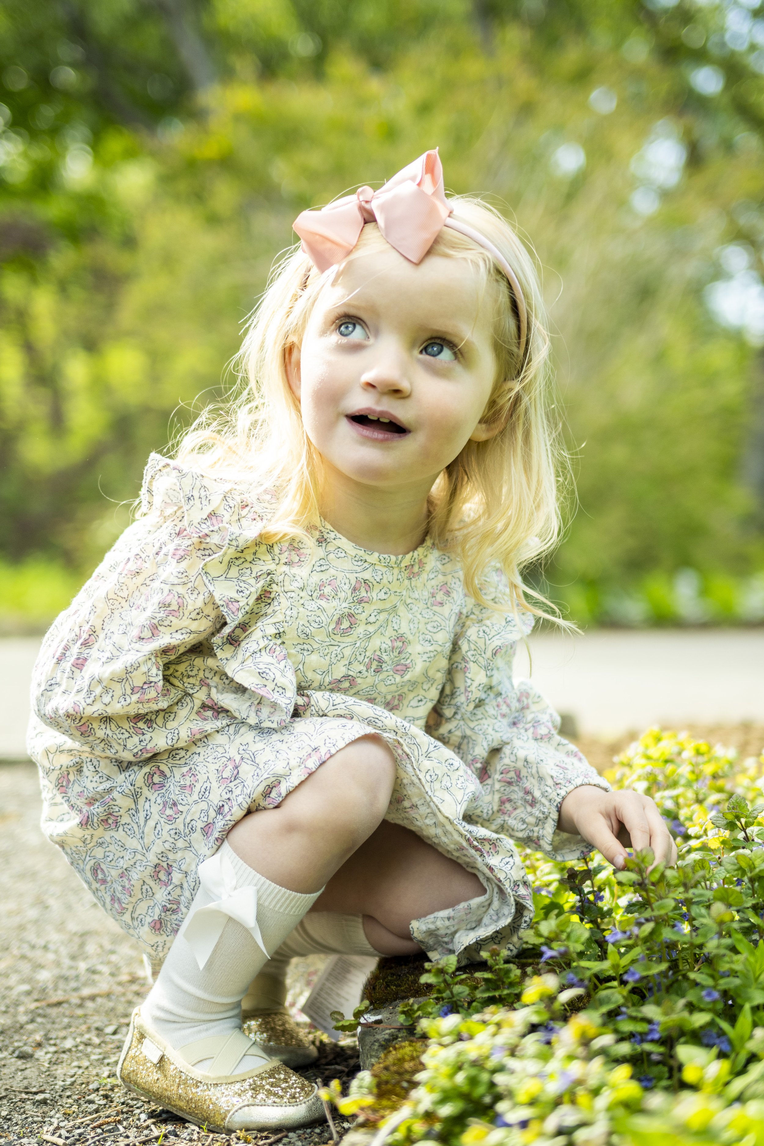 A young girl with blonde hair, dressed in a floral dress and white socks with a bow, is crouching outdoors in a garden surrounded by green foliage and colorful flowers, looking up thoughtfully.