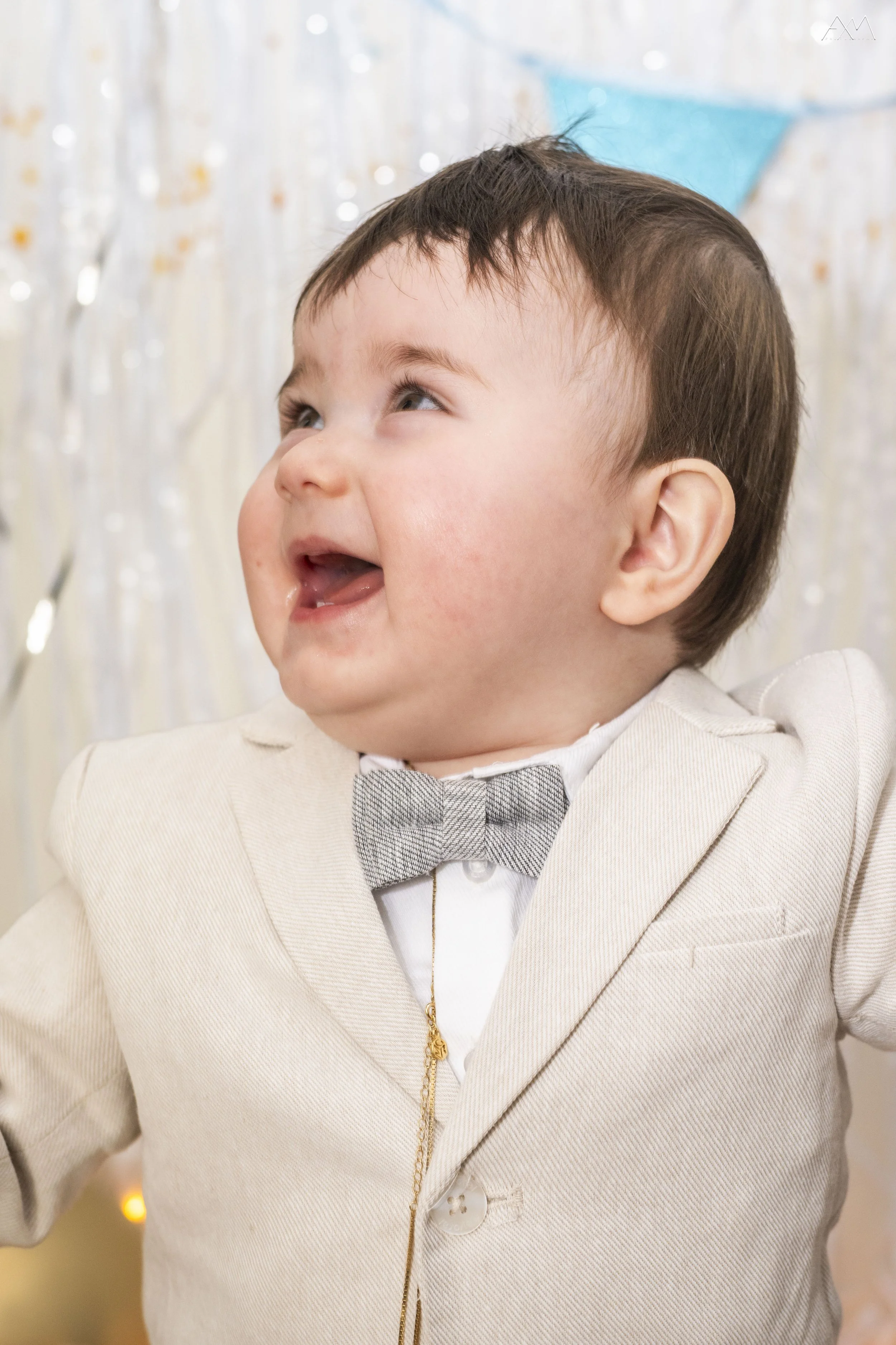 Portrait of a young child dressed in a beige suit with a plaid bow tie, smiling and looking up, against a festive background with blurred lights and decorations.