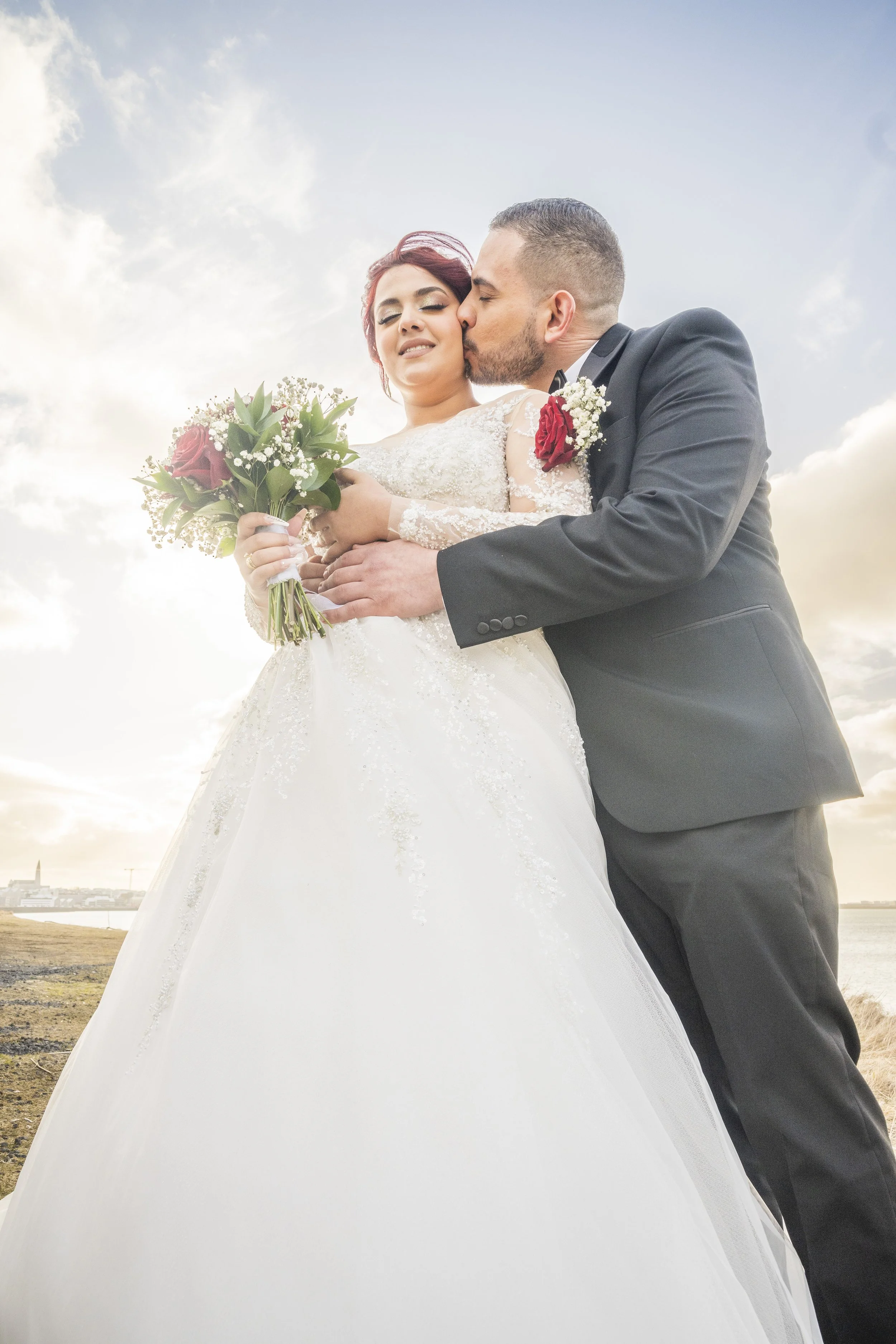 A bride and groom sharing a kiss outdoors during their wedding, with the bride holding a bouquet of red roses and greenery, sky with clouds in the background.