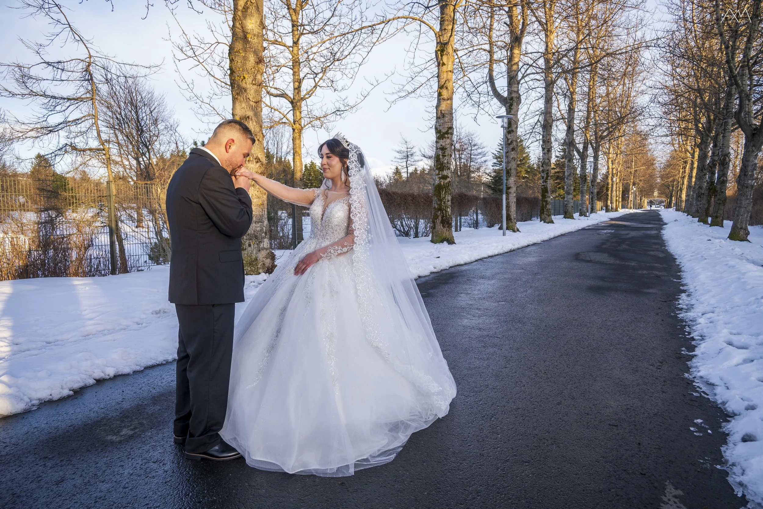 A bride and groom standing on a snow-covered road lined with tall leafless trees, with the groom kissing the bride's hand during sunset.