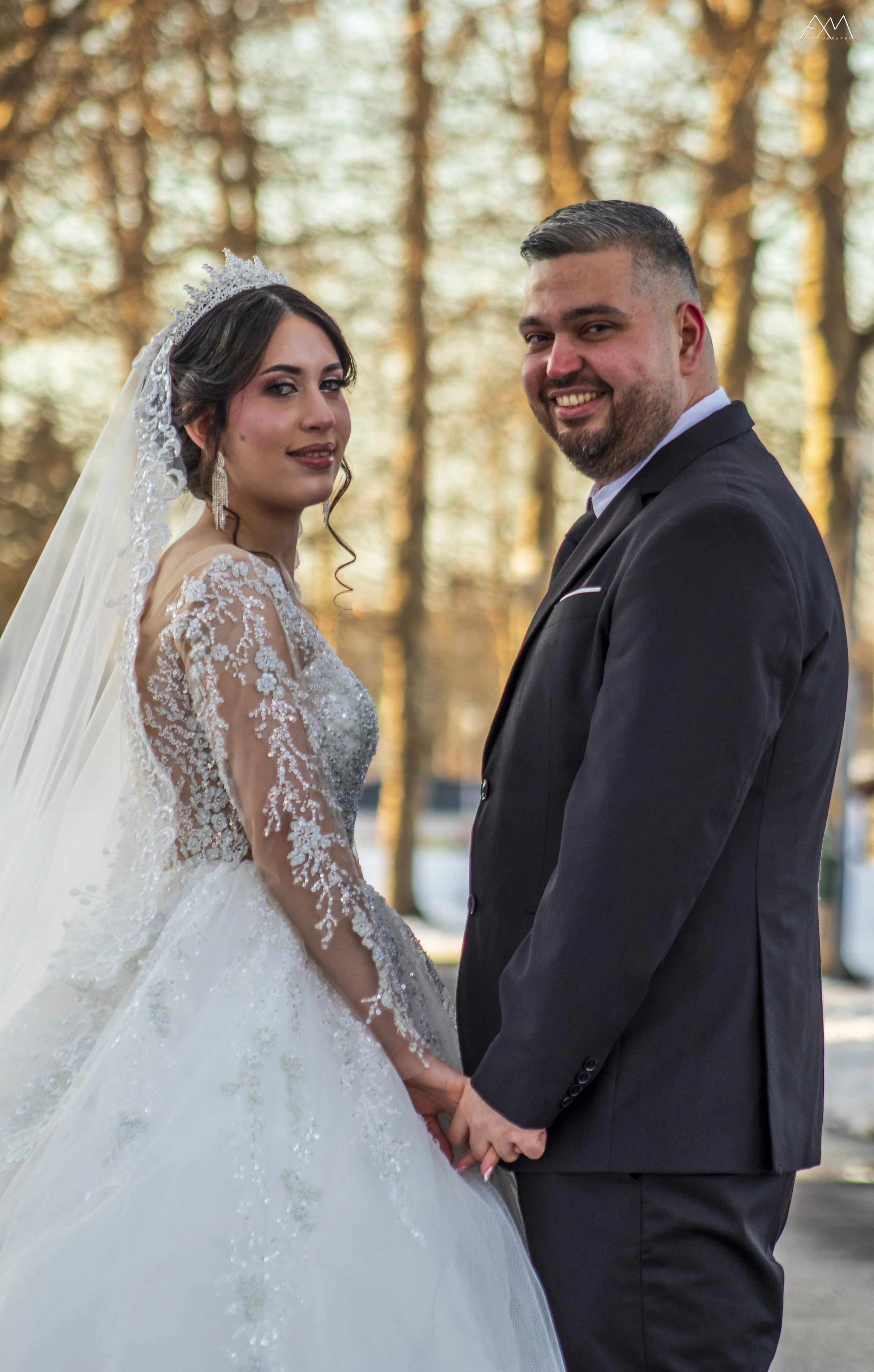 A bride and groom holding hands outdoors during a wedding, with the bride in a lace wedding gown and tiara, and the groom in a black suit, standing in front of trees with sunlight filtering through.