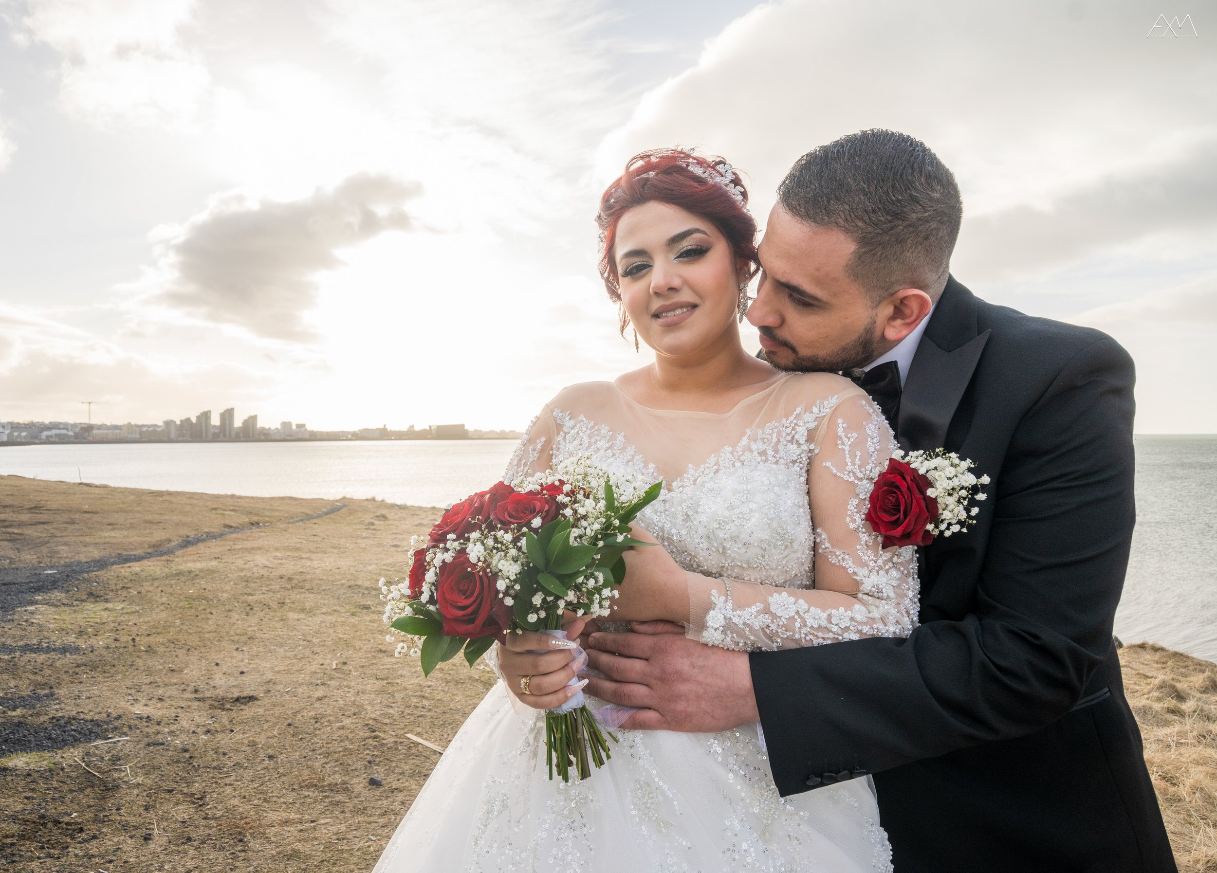 Bride and groom standing on a grassy shoreline by the water, with the bride holding a bouquet of red roses and the groom embracing her from the side.