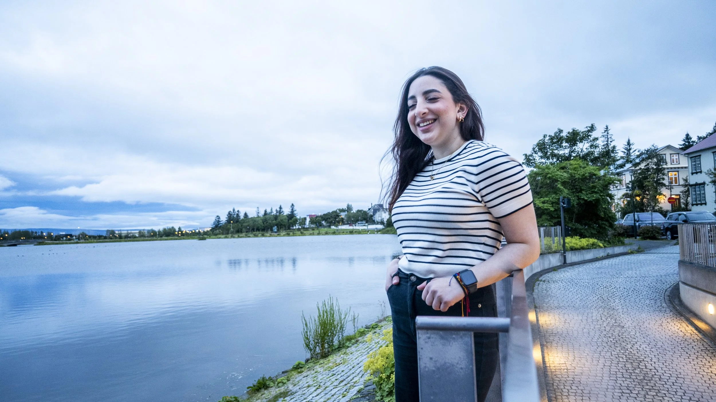 Smiling young woman with dark hair wearing a striped shirt, standing near a river with houses and trees in the background during evening or early night.