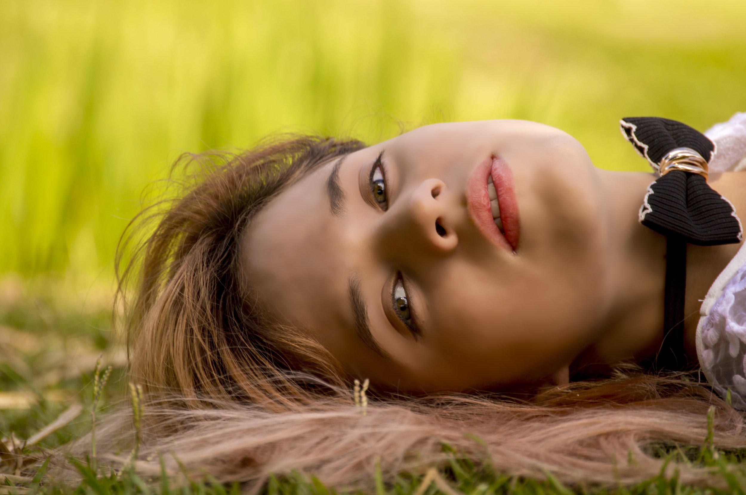 A woman with long hair lying on grass in a natural setting, looking up with a relaxed expression.