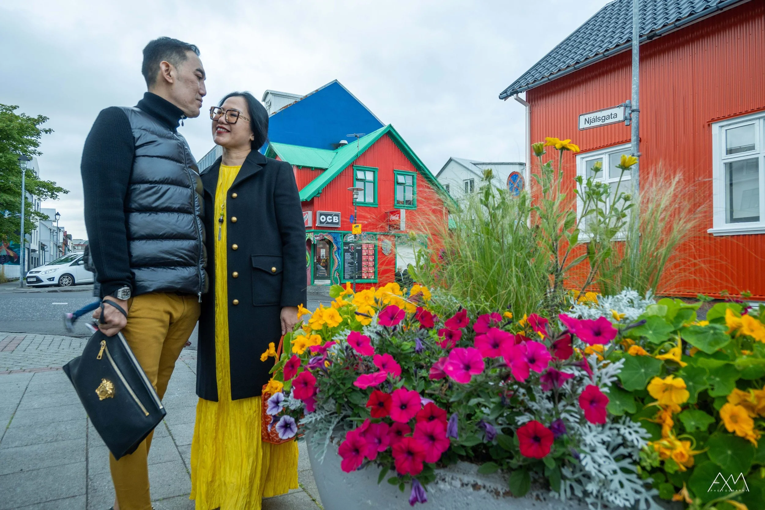 A man and woman stand close together and smile on a city street with colorful buildings, flowers, and a street sign reading 'Njálsgata'.