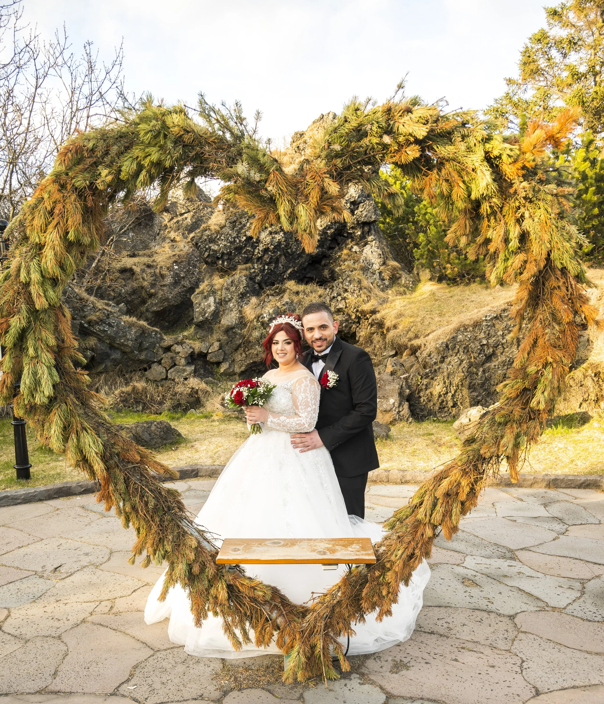 A newlywed couple stands in front of a rocky outdoor setting, framed by a heart-shaped arch decorated with green and brown pine branches.