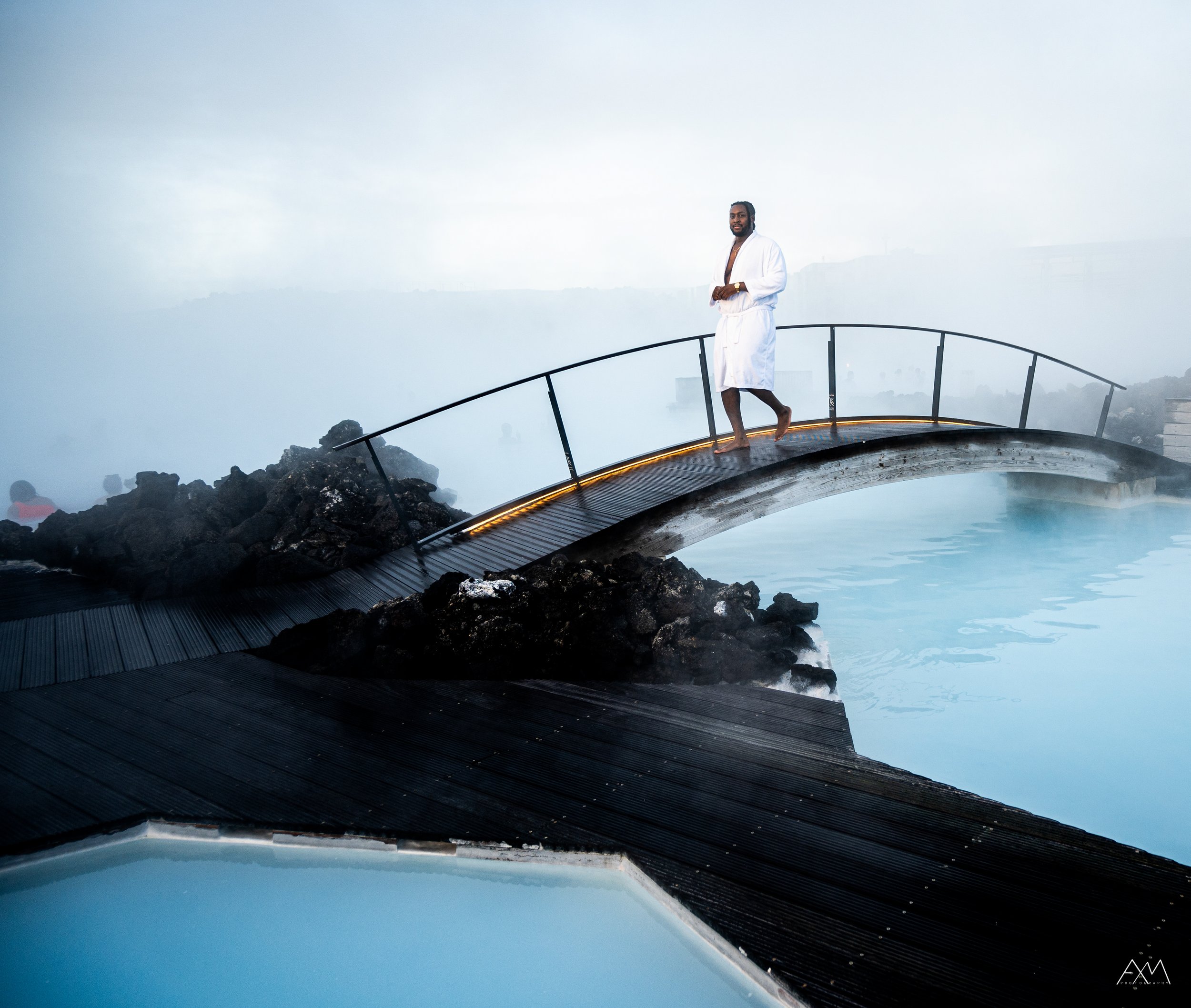 A man in a white robe standing on a small wooden bridge over a hot spring or blue lagoon in Iceland with steam rising around. The background is foggy and misty.