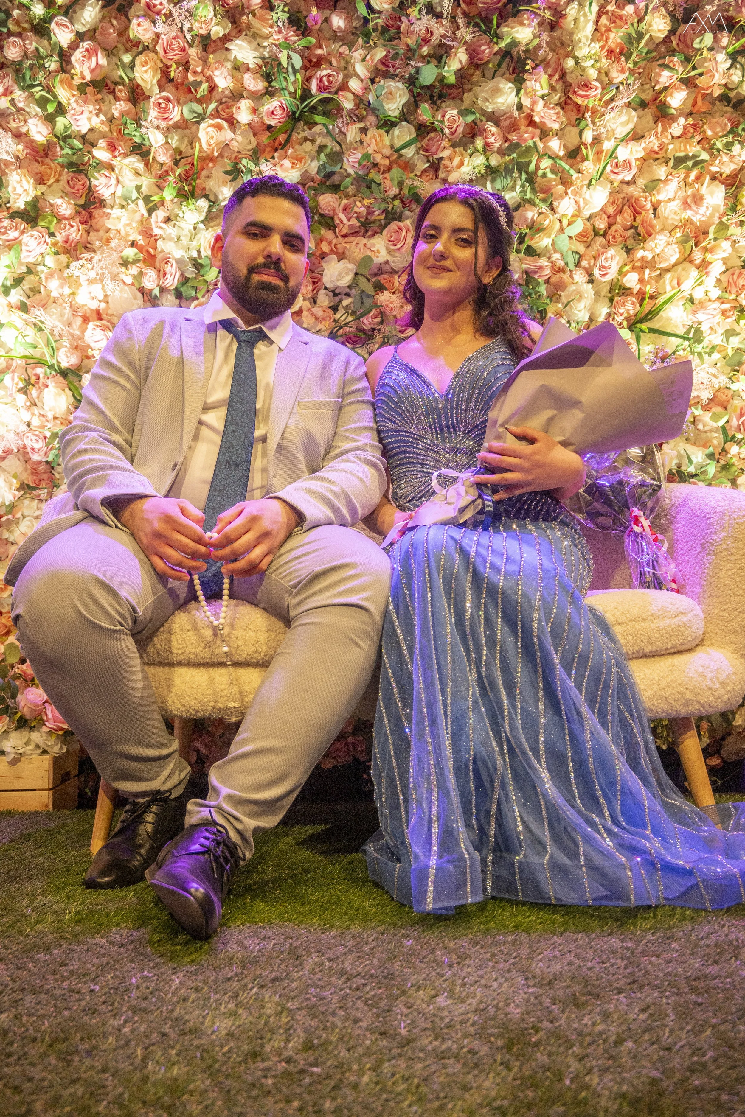 Man and woman sitting on a striped couch in front of an elaborate flower wall, with the woman holding a bouquet of flowers.