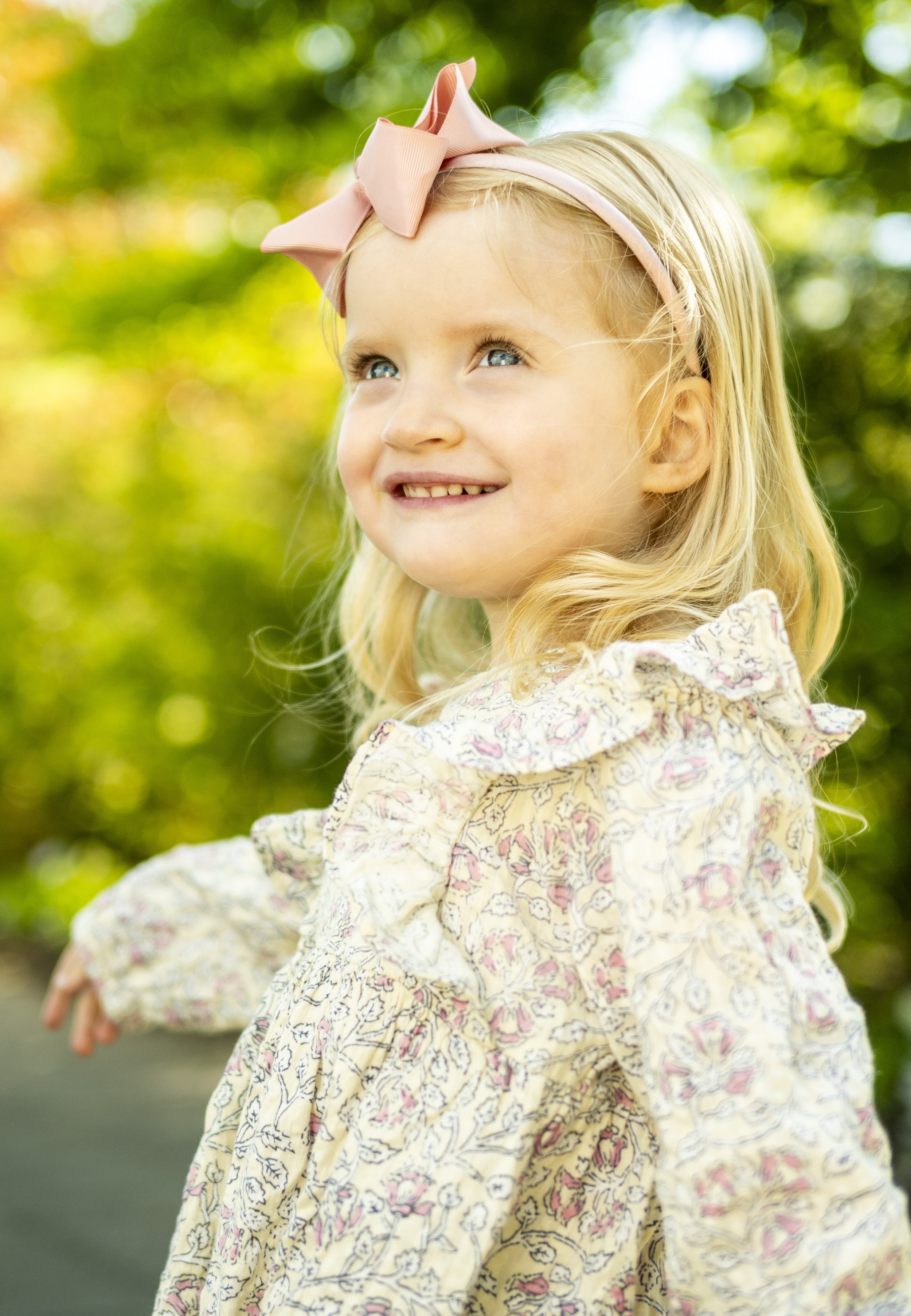 A young girl with blonde hair, blue eyes, and a pink bow, smiling outdoors with green foliage in the background.