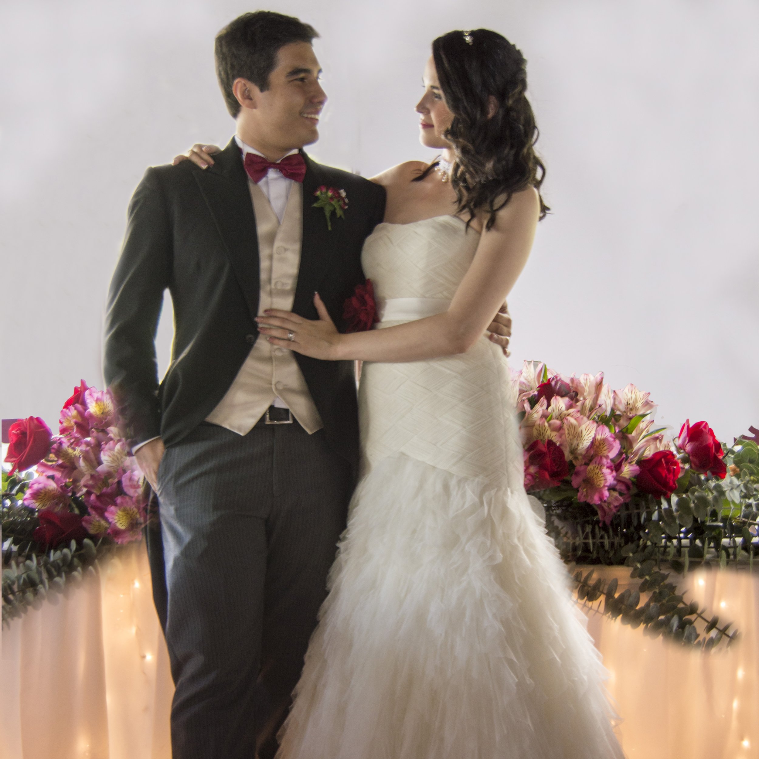 A bride and groom at their wedding, standing close and looking into each other's eyes, with floral arrangements in the background.