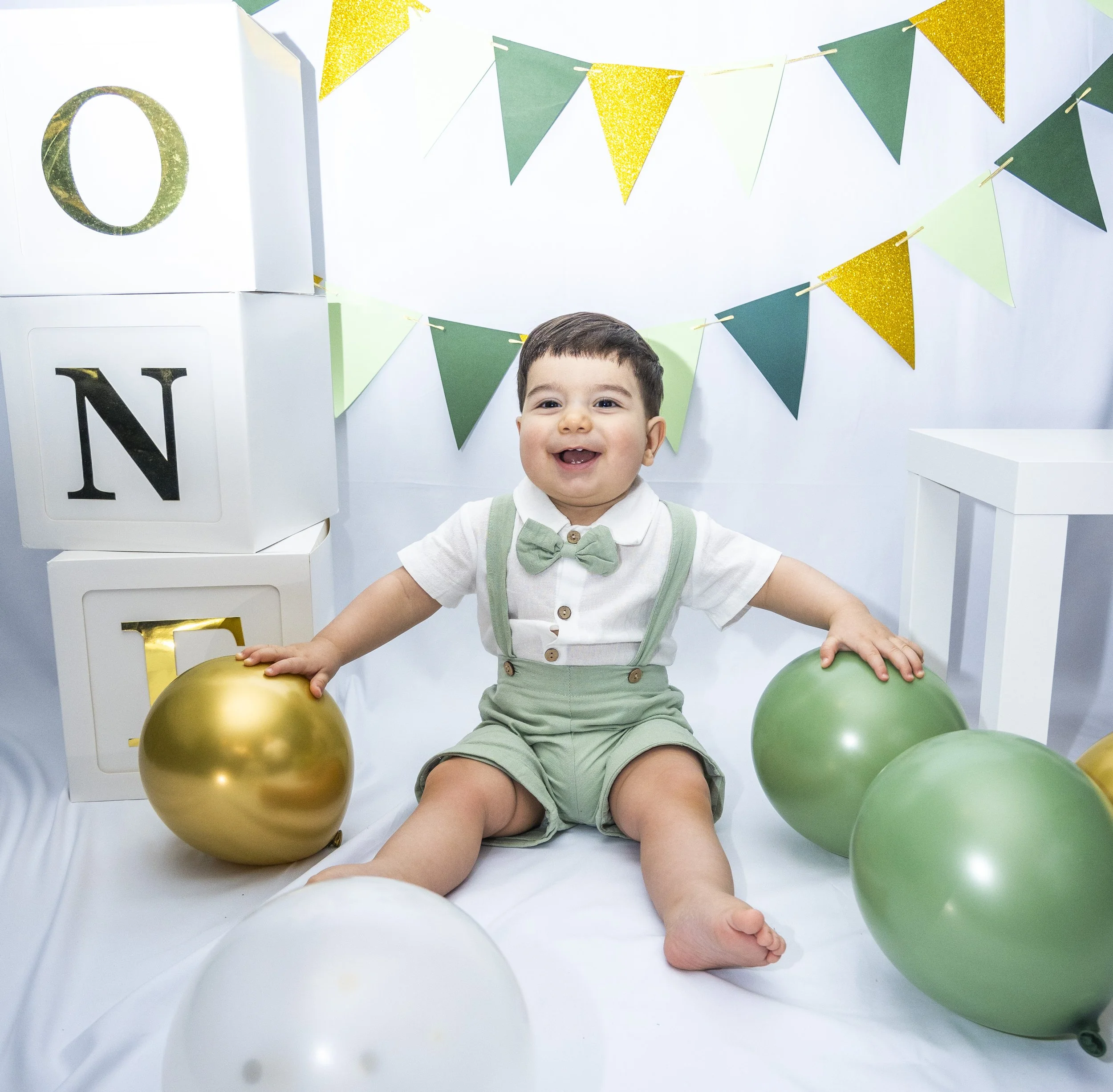 A young boy smiling at the camera with balloons and green and gold bunting decoration in the background.