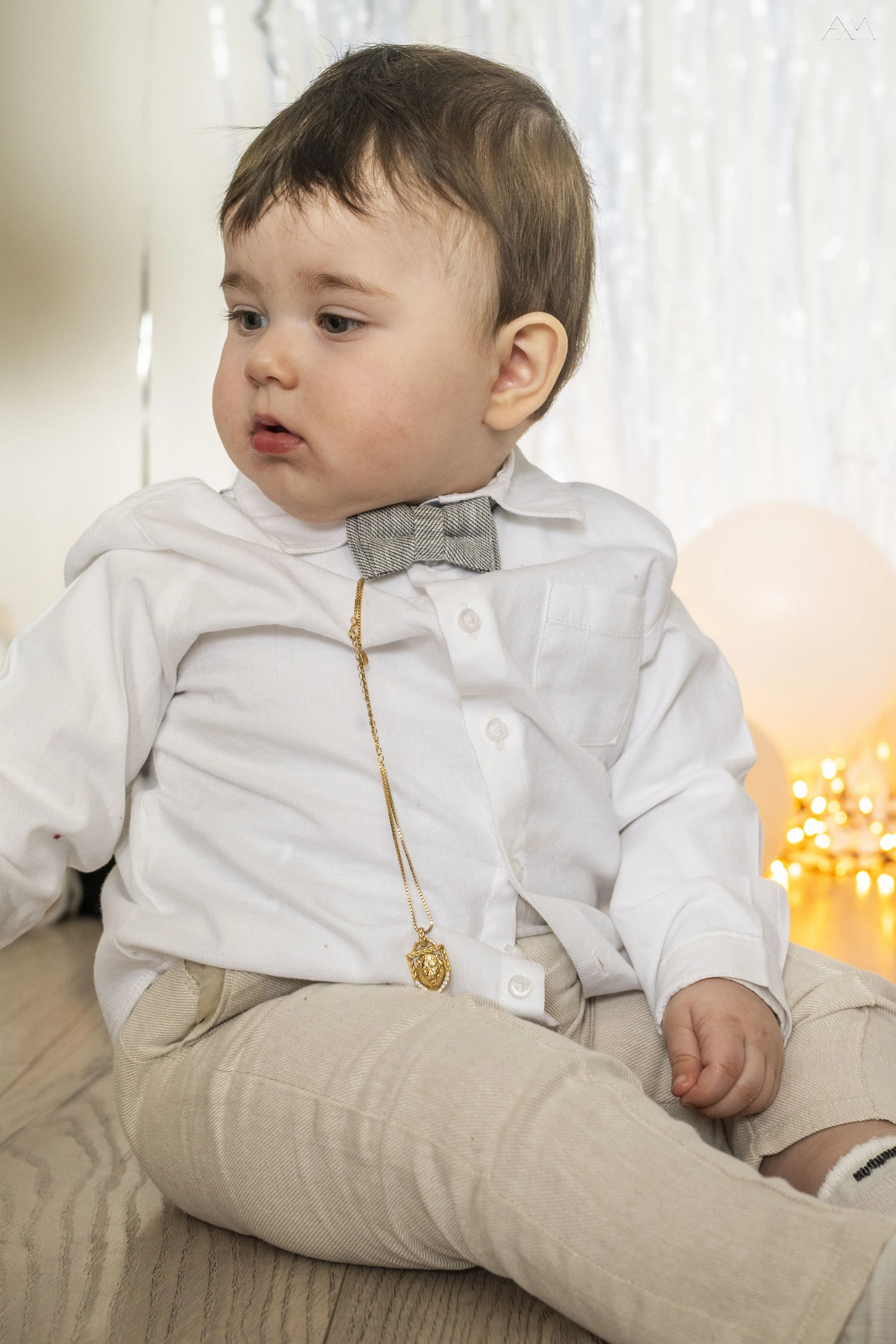 Young boy sitting on wooden floor wearing a white shirt, beige pants, and a gray bow tie, with a gold chain and pendant, looking to the side with a neutral expression.