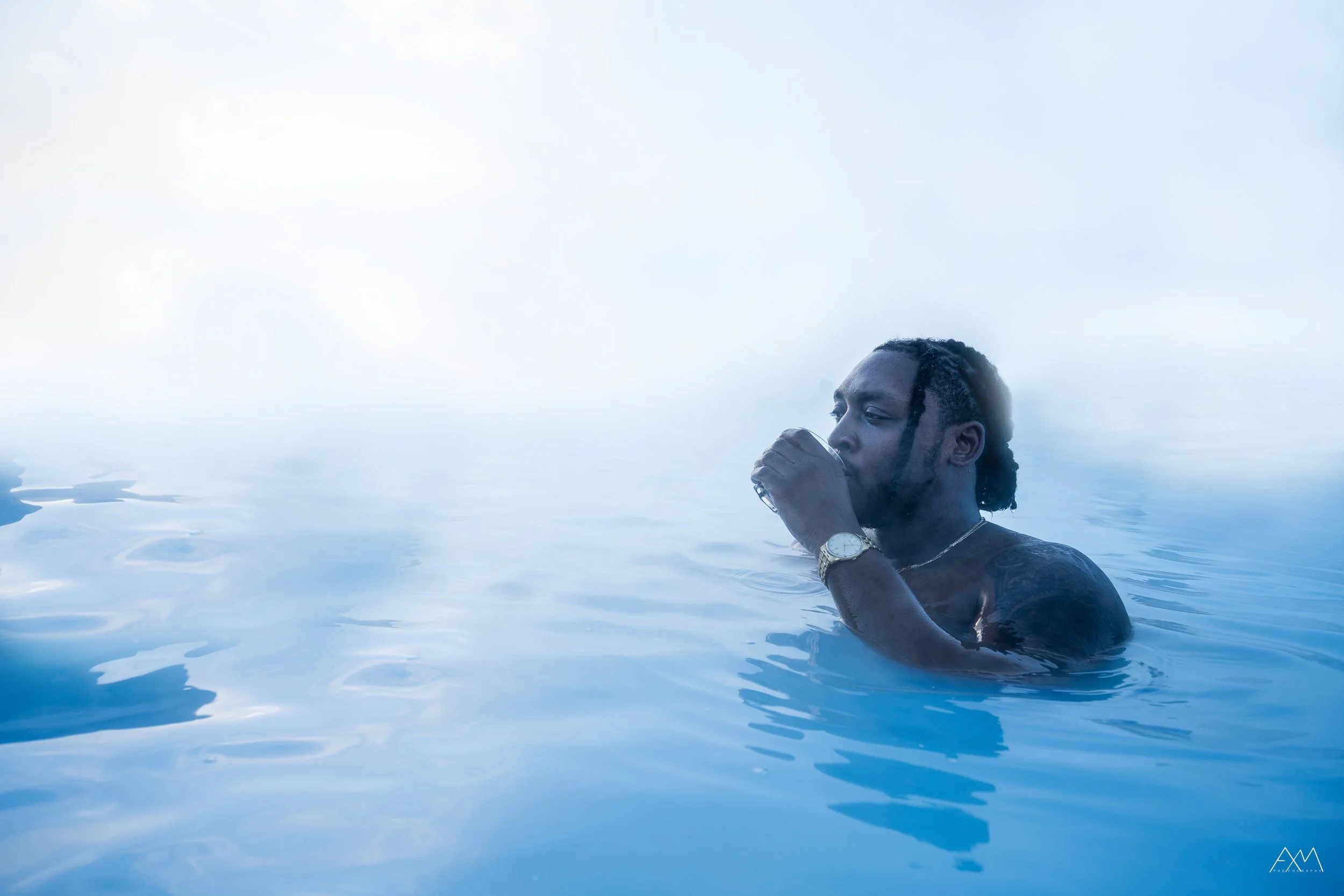 Man with dreadlocks and watch drinking water or beverage in a misty, blue-toned pool or hot spring.