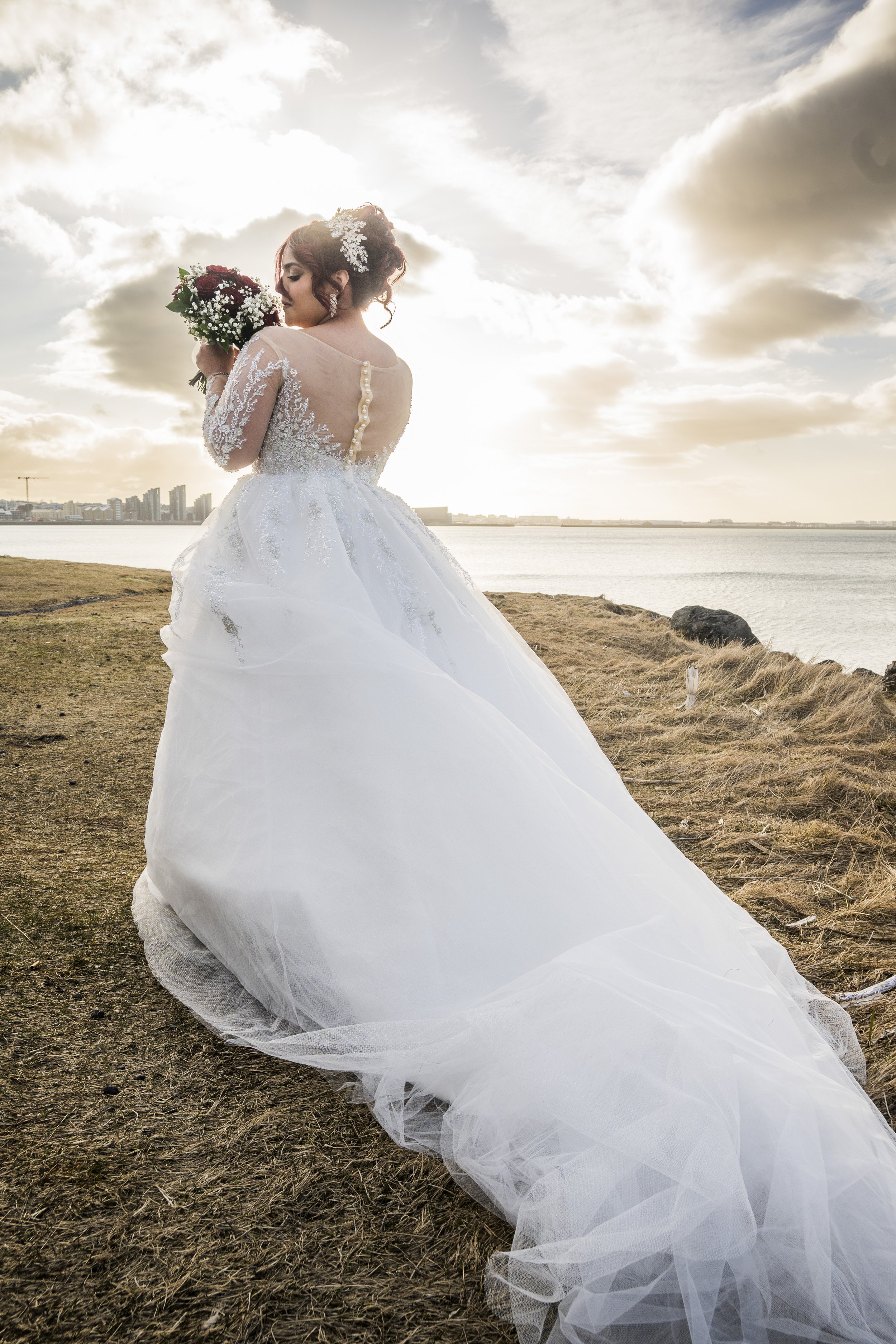 A woman in a wedding dress holding a bouquet of flowers by a body of water during sunset.
