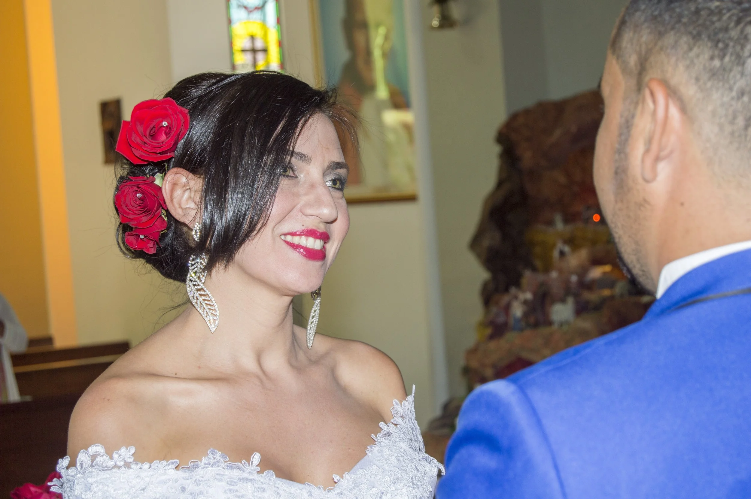 A woman with dark hair adorned with red roses is smiling and looking at a man in a blue suit during a wedding ceremony inside a church with stained glass windows and religious artwork.