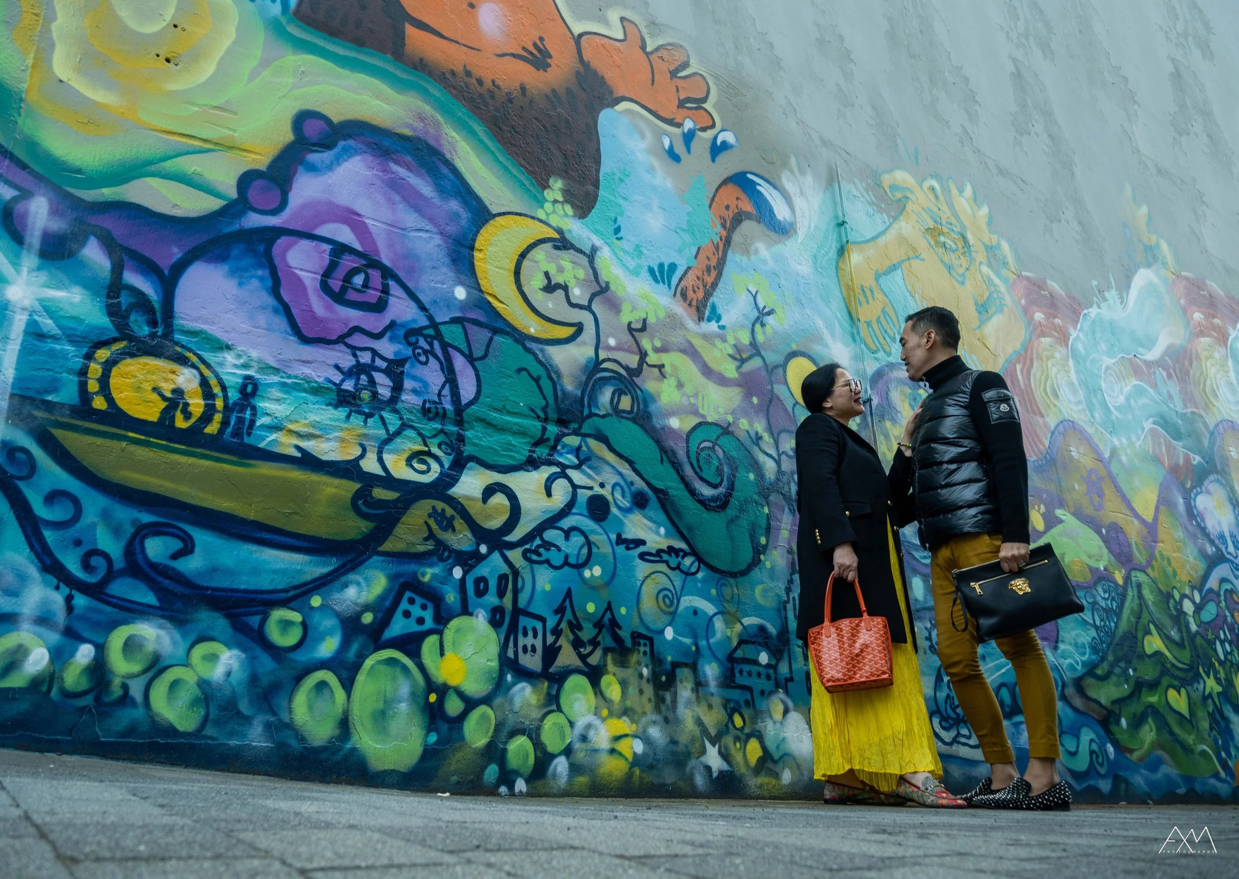 Two people standing and talking in front of a colorful graffiti mural on a wall. The woman is wearing glasses, a dark jacket, a yellow skirt, and holding a red handbag. The man is wearing a black puffer jacket, tan pants, black shoes with white dots,