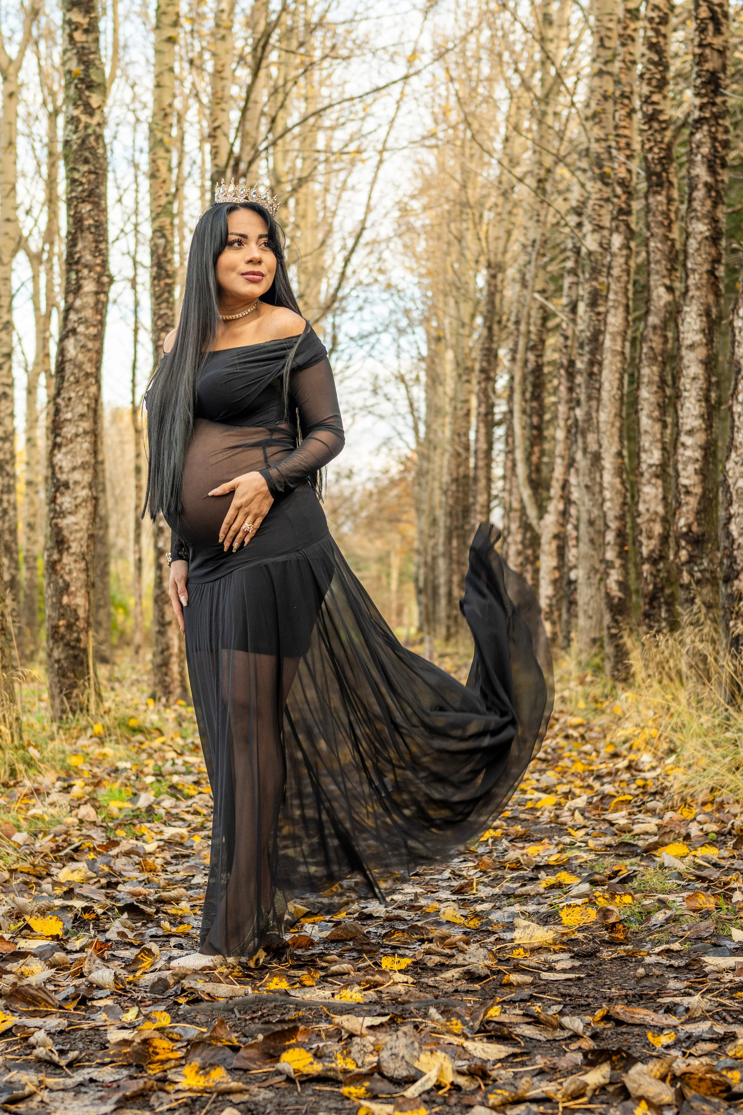 A pregnant woman in a sheer black dress with an off-the-shoulder style, wearing a crown, standing in a forest with autumn leaves on the ground, looking to her left with a serene expression.