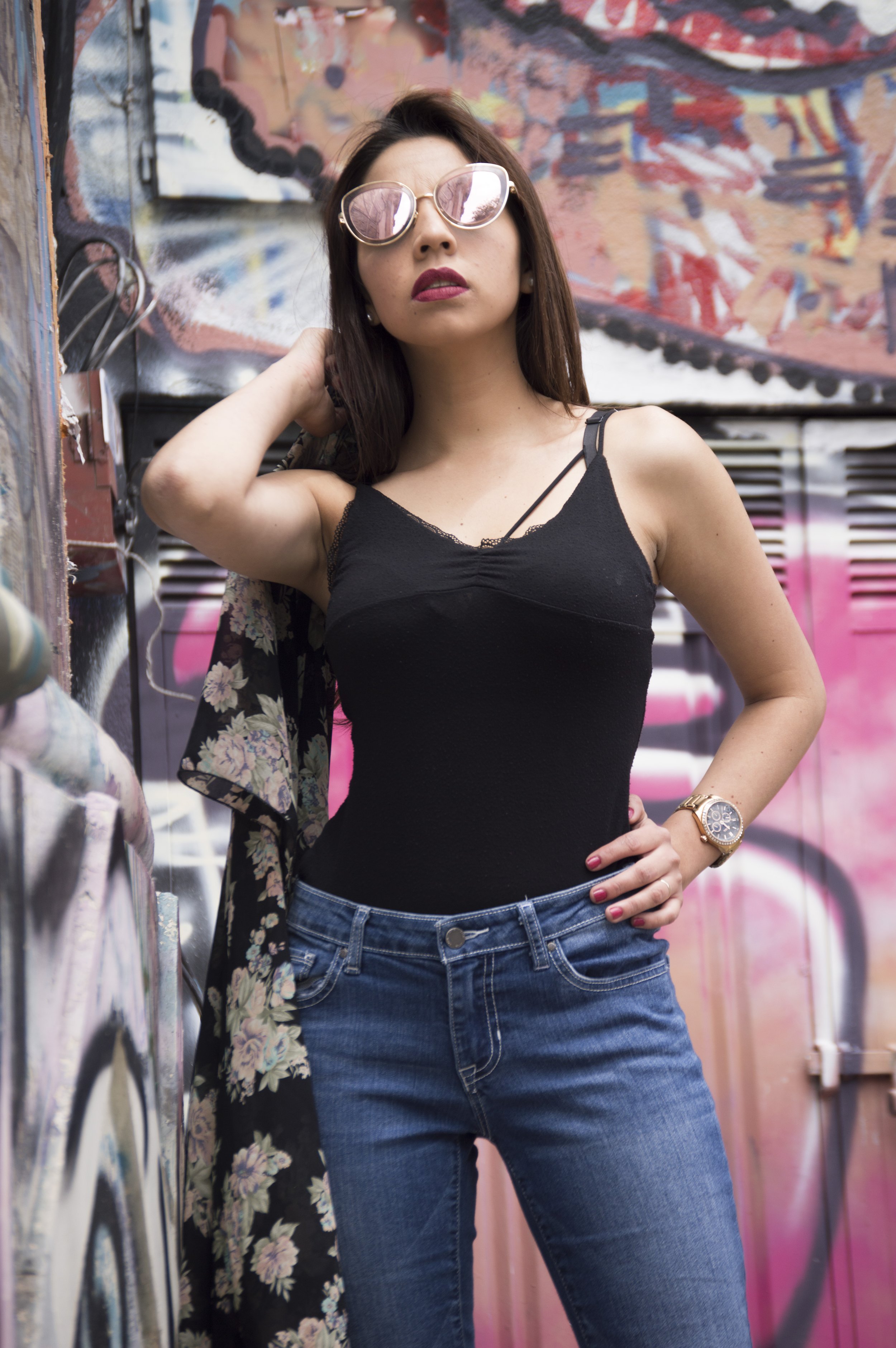 A young woman with long brown hair sunglasses, dressed in a black tank top and blue jeans, standing against a colorful graffiti wall.